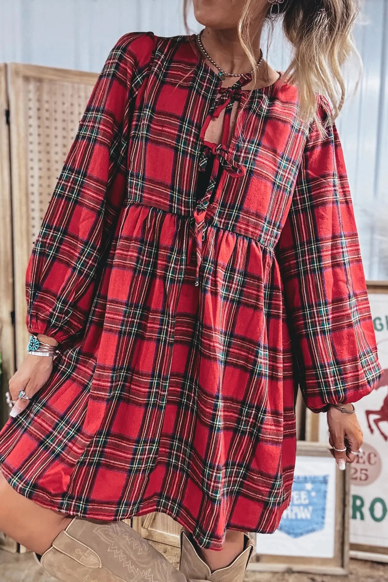 A woman wearing a red plaid long-sleeve dress with a tie-front detail and gathered waist, paired with tan boots in a rustic indoor setting.