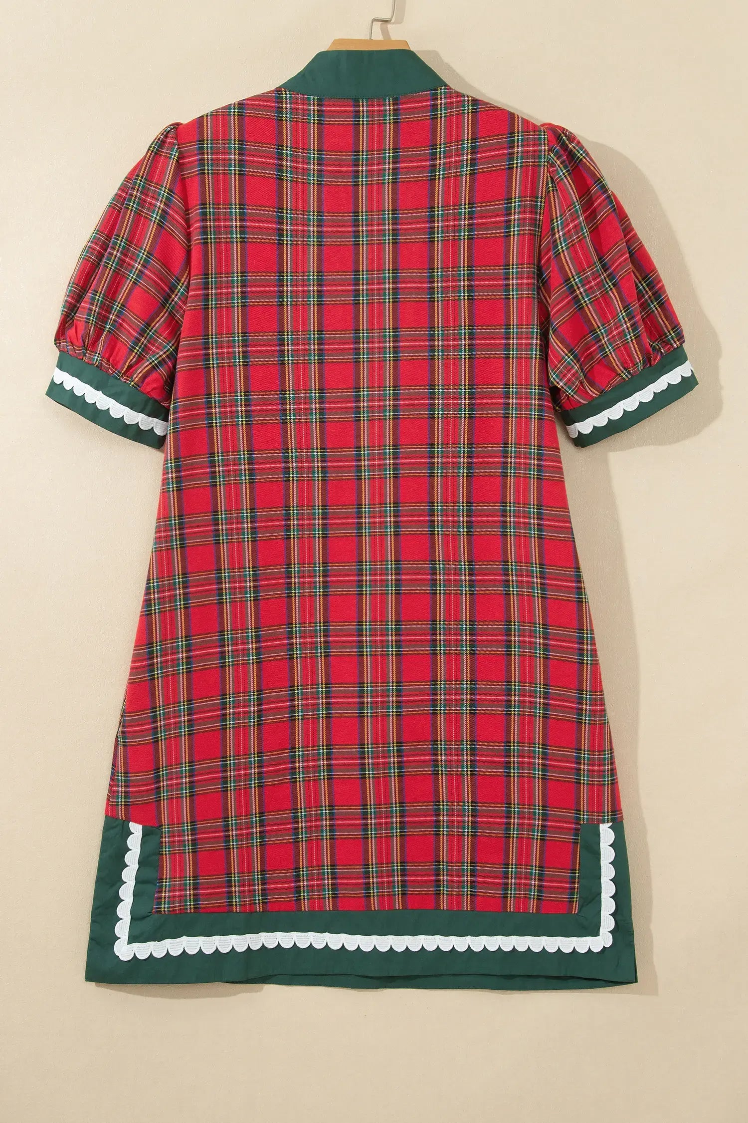 A red and green plaid short-sleeve dress with dark green trim and white scalloped edges, shown hanging against a plain beige wall.