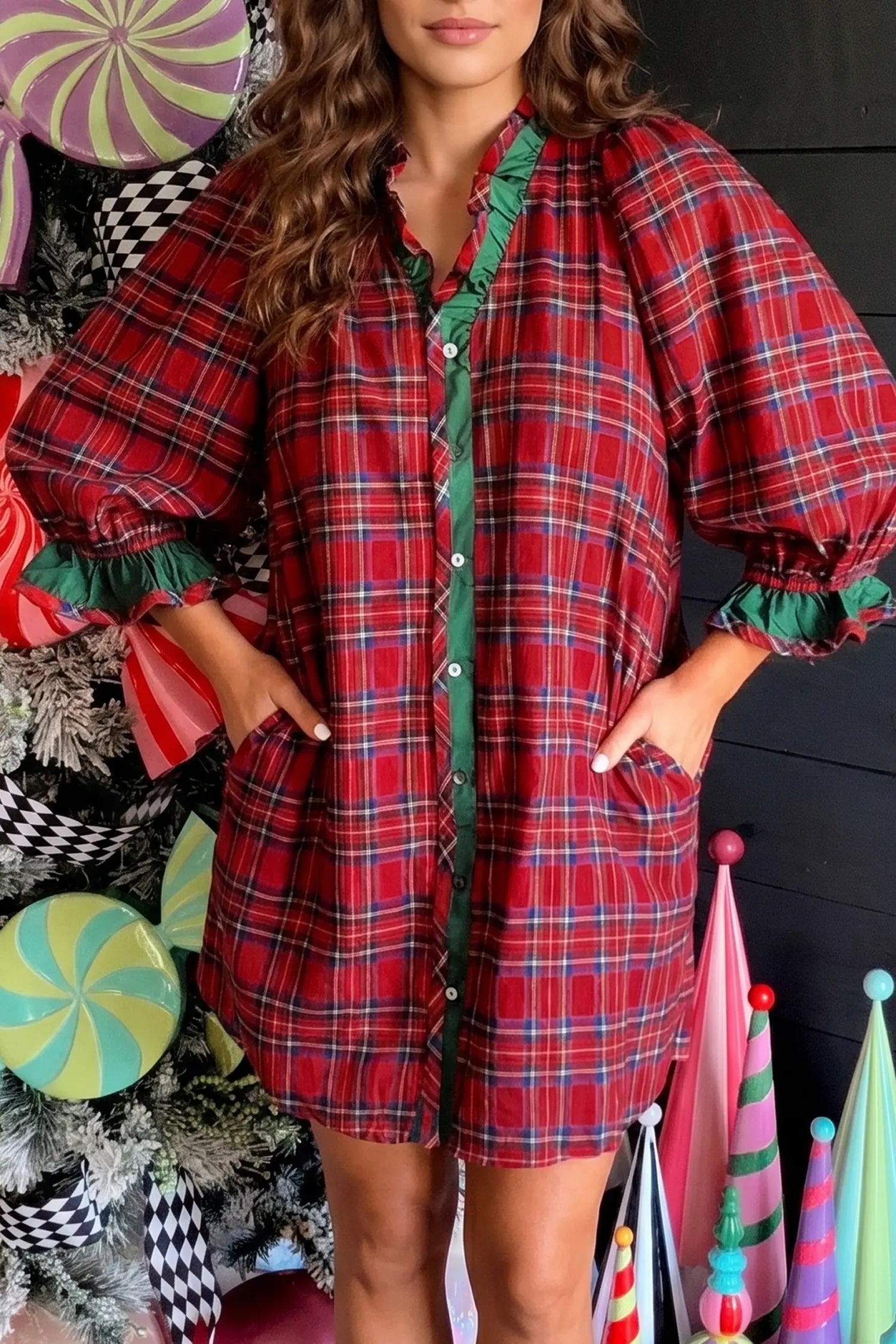 A woman wearing a red and green plaid button-up shirt dress with ruffled trim, posed in front of a decorated holiday background with candy decorations and party hats.
