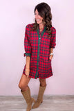 A woman modeling a red and green plaid mini shirt dress with brown suede boots, standing against a light pink wall.