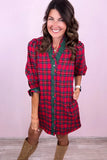 A woman wearing a red and green plaid button-down shirt dress with three-quarter sleeves and brown boots, standing against a light pink wall.