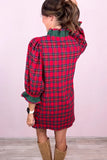 A woman shown from the back wearing a red and green plaid puff-sleeve dress with a green ruffled collar and cuffs, paired with tan boots, against a light pink wall.