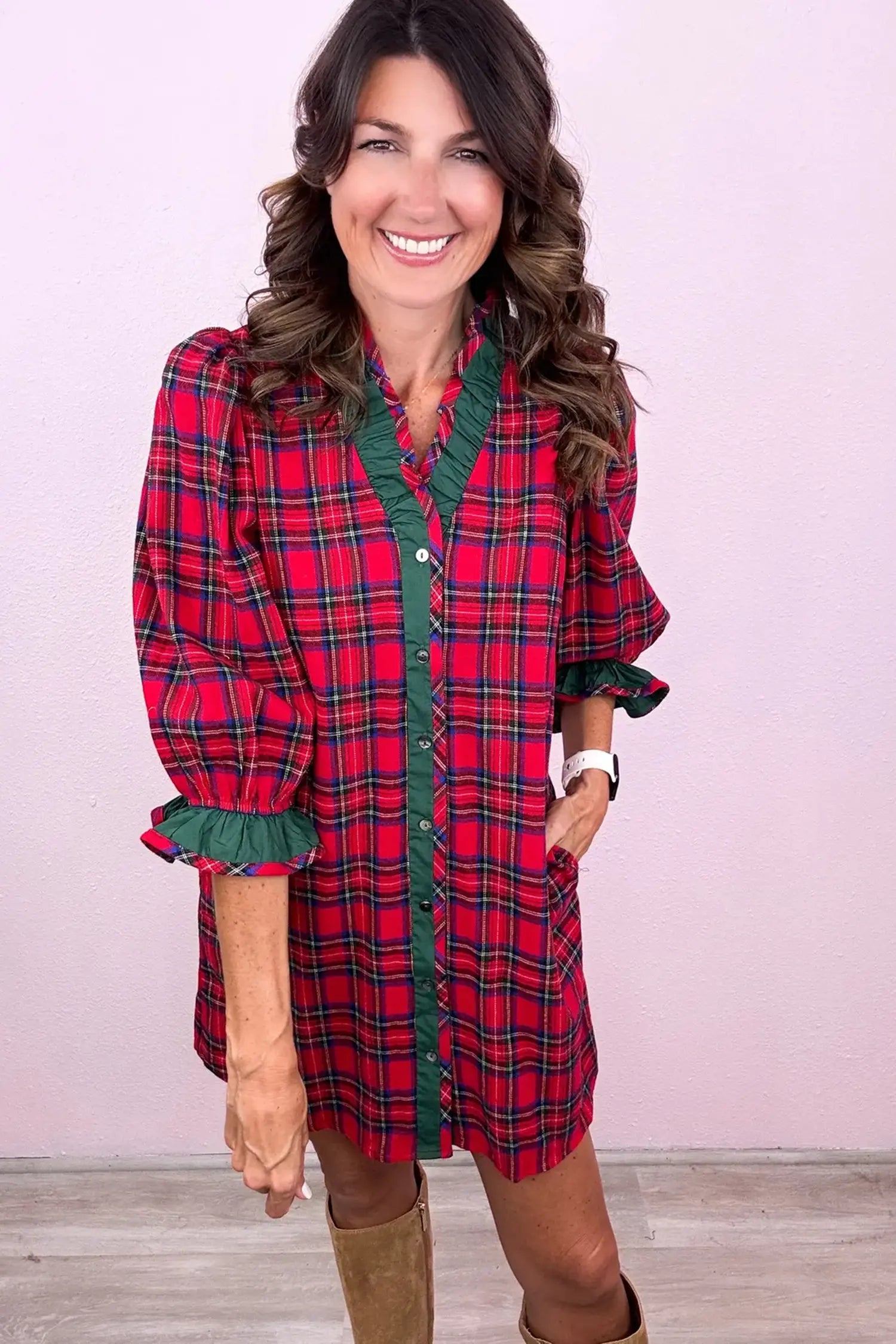 A woman wearing a red and green plaid button-down shirt dress with ruffle trim, standing against a light pink wall.