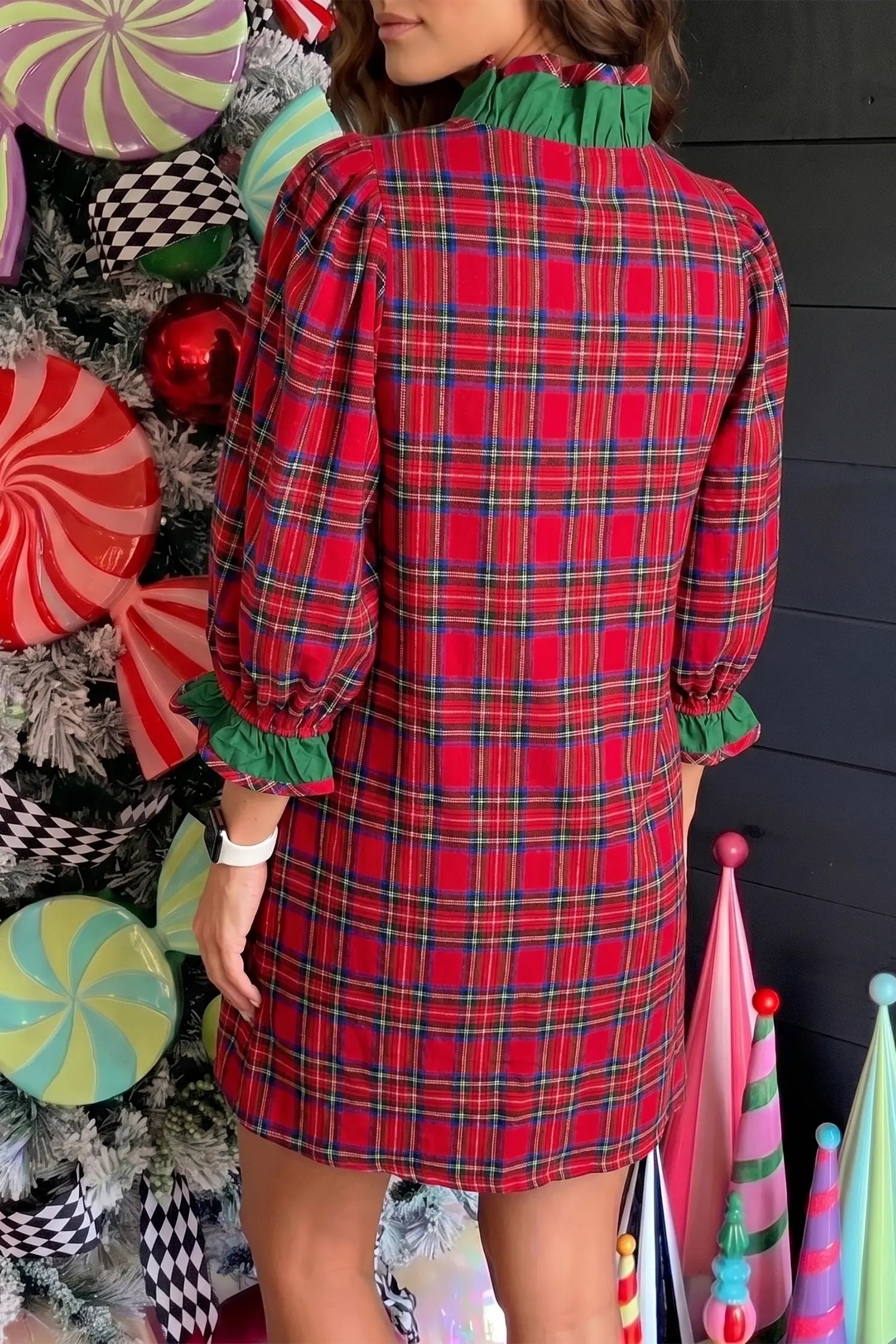A woman wearing a red and green plaid long-sleeve dress with a green ruffled collar and ruffled cuffs, photographed from the back in a festive holiday-themed setting.