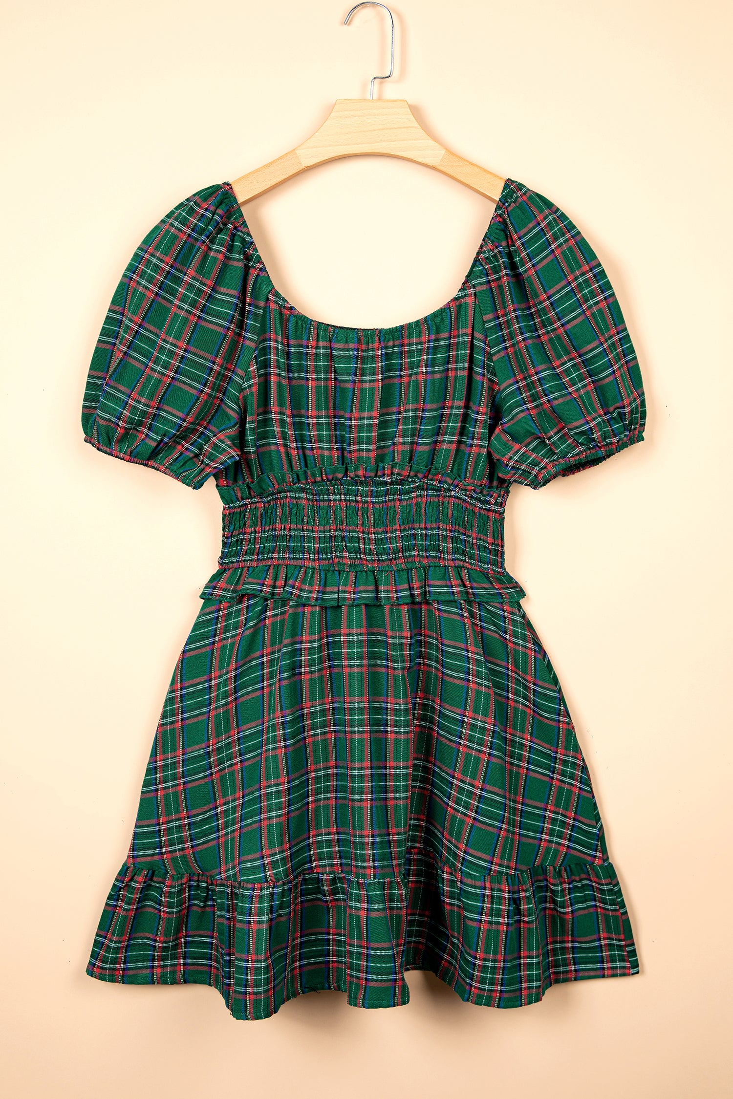 A green plaid mini dress with puff sleeves and a smocked waist, hanging on a wooden hanger against a solid beige background.
