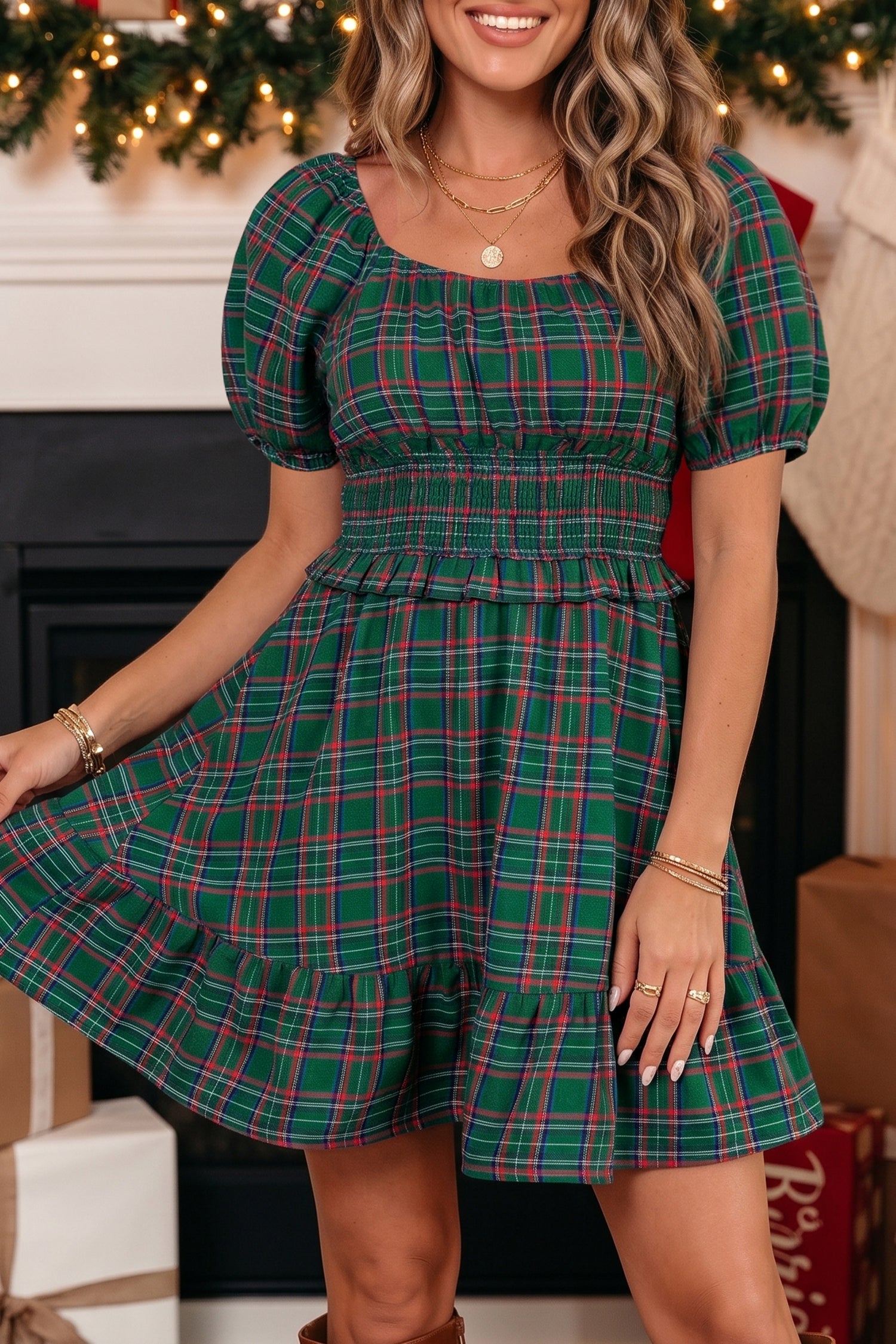 A woman wearing a festive green plaid dress with puff sleeves and a smocked waist, posed in front of a decorated fireplace.