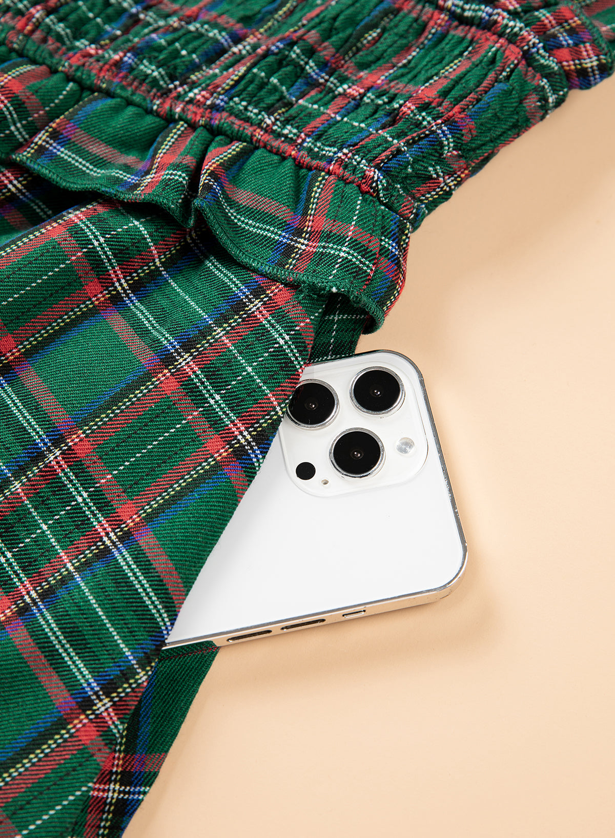 A close-up of green plaid fabric with red, white, and blue tartan lines, draped over a light beige surface with a white smartphone partially visible underneath.