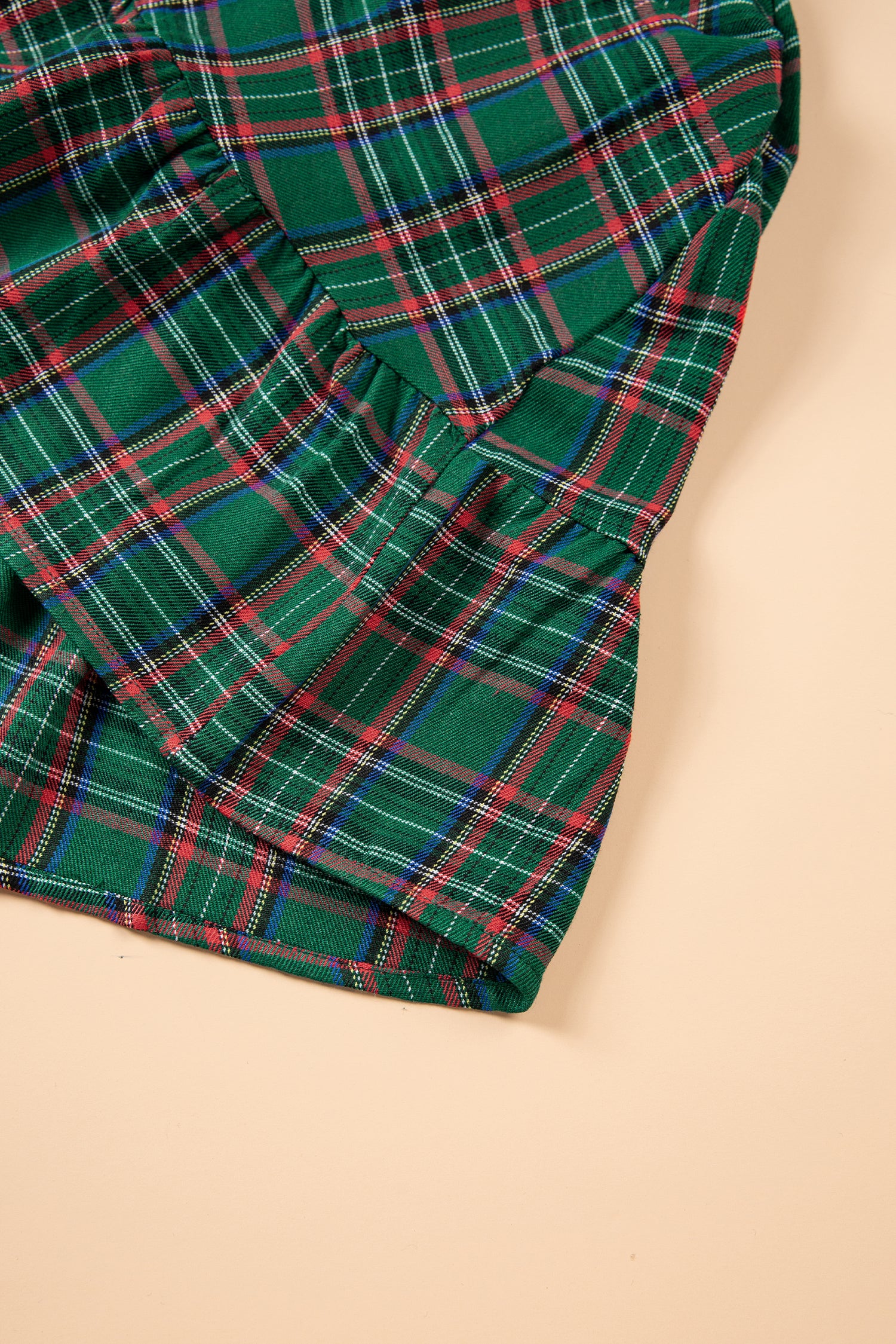 A close-up of a green tartan plaid fabric with red, white, and blue intersecting stripes, folded to show its texture and pattern against a solid beige background.
