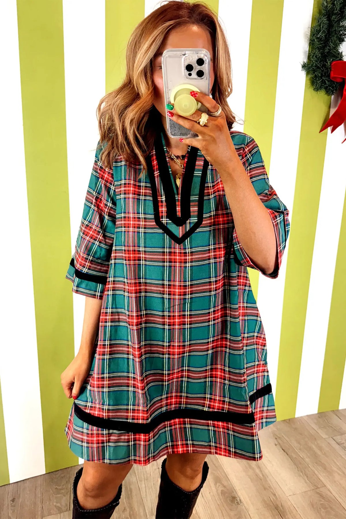 A woman takes a mirror selfie wearing a festive plaid tunic dress with red, green, and white stripes and black trim, set against a lime green and white striped wall.