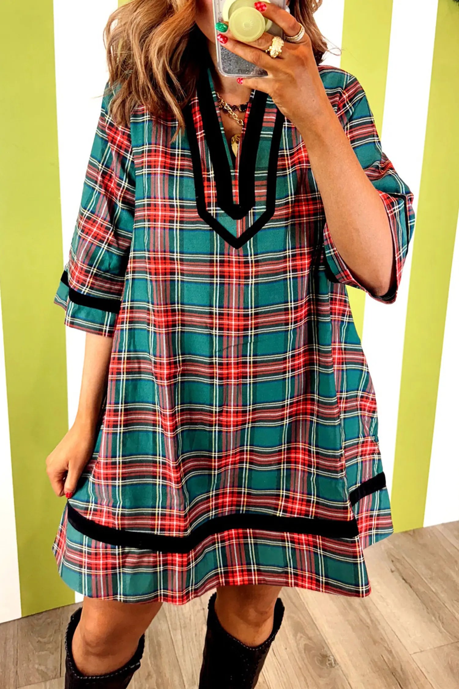 A woman takes a mirror selfie wearing a short-sleeve plaid tunic dress with a teal, red, and white pattern and black trim. She is wearing black boots and standing in front of a green and white striped wall.