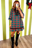 A woman takes a mirror selfie wearing a short-sleeve plaid shirt dress in green, red, and white with black trim, paired with black knee-high boots, set against a green and white striped wall.
