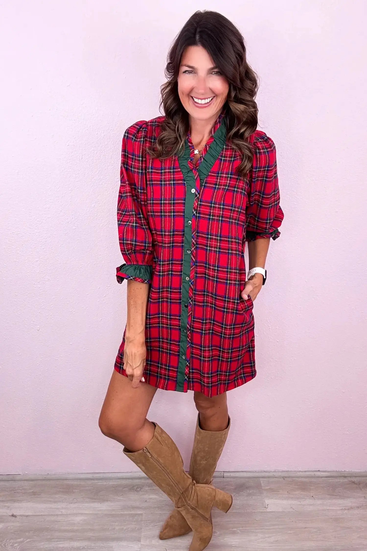 A woman modeling a red and green plaid mini shirt dress with brown suede knee-high boots, standing against a light pink wall.