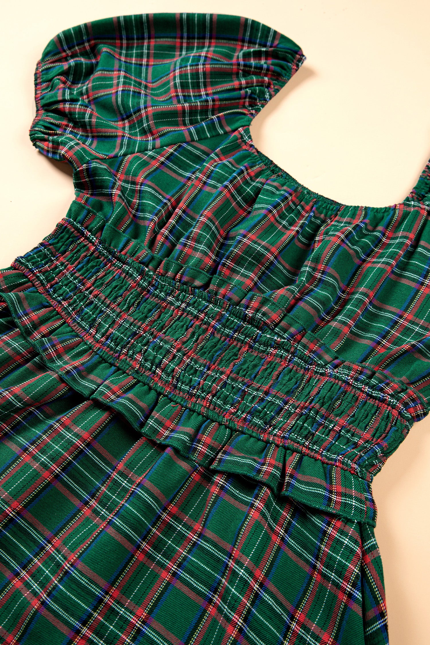 A close-up of a green plaid dress with a smocked bodice and ruffled straps, laid flat on a light beige background.