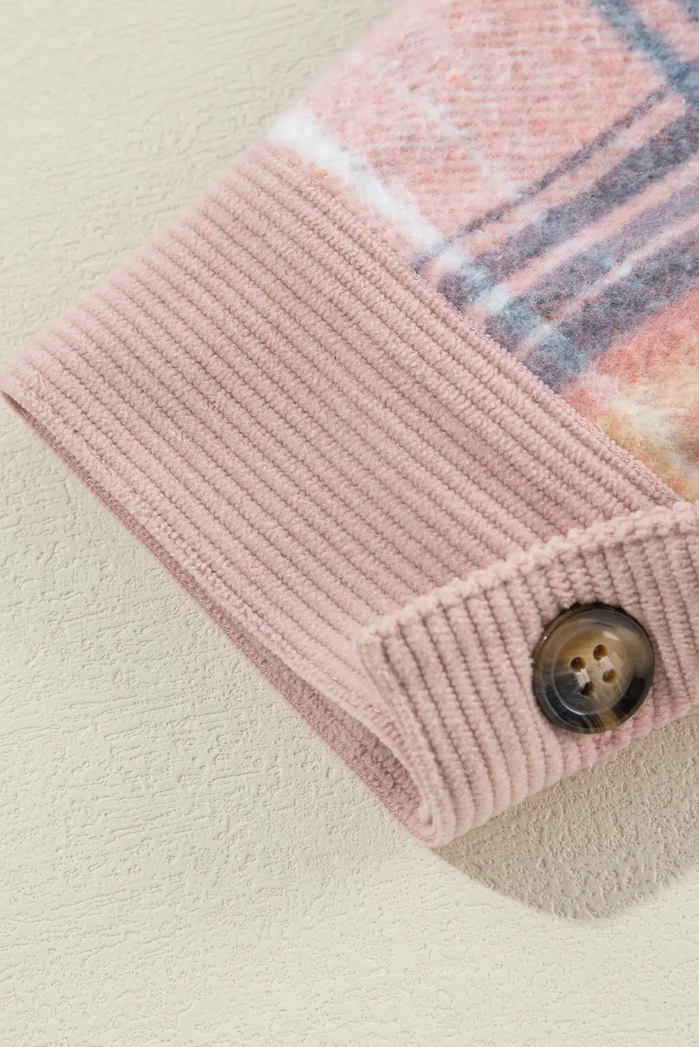 Close-up of a folded pink ribbed knit garment, likely a sweater or cardigan, showing a plaid pattern in pink, blue, and white at the cuff, with a brown button closure on a textured cream background.