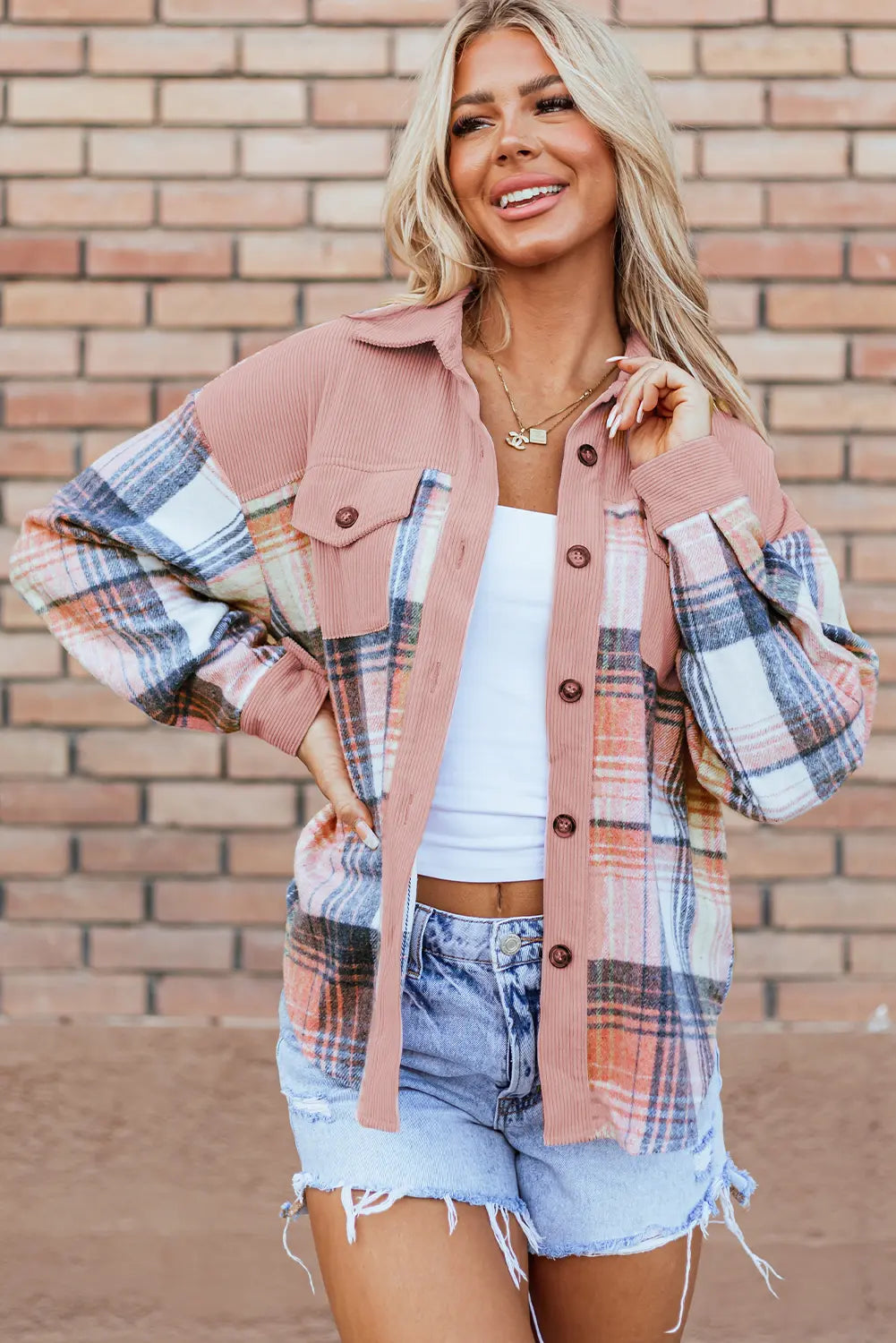 A woman models a dusty pink and white plaid button-up shirt over a white tank top and denim shorts, standing against a brick wall background.