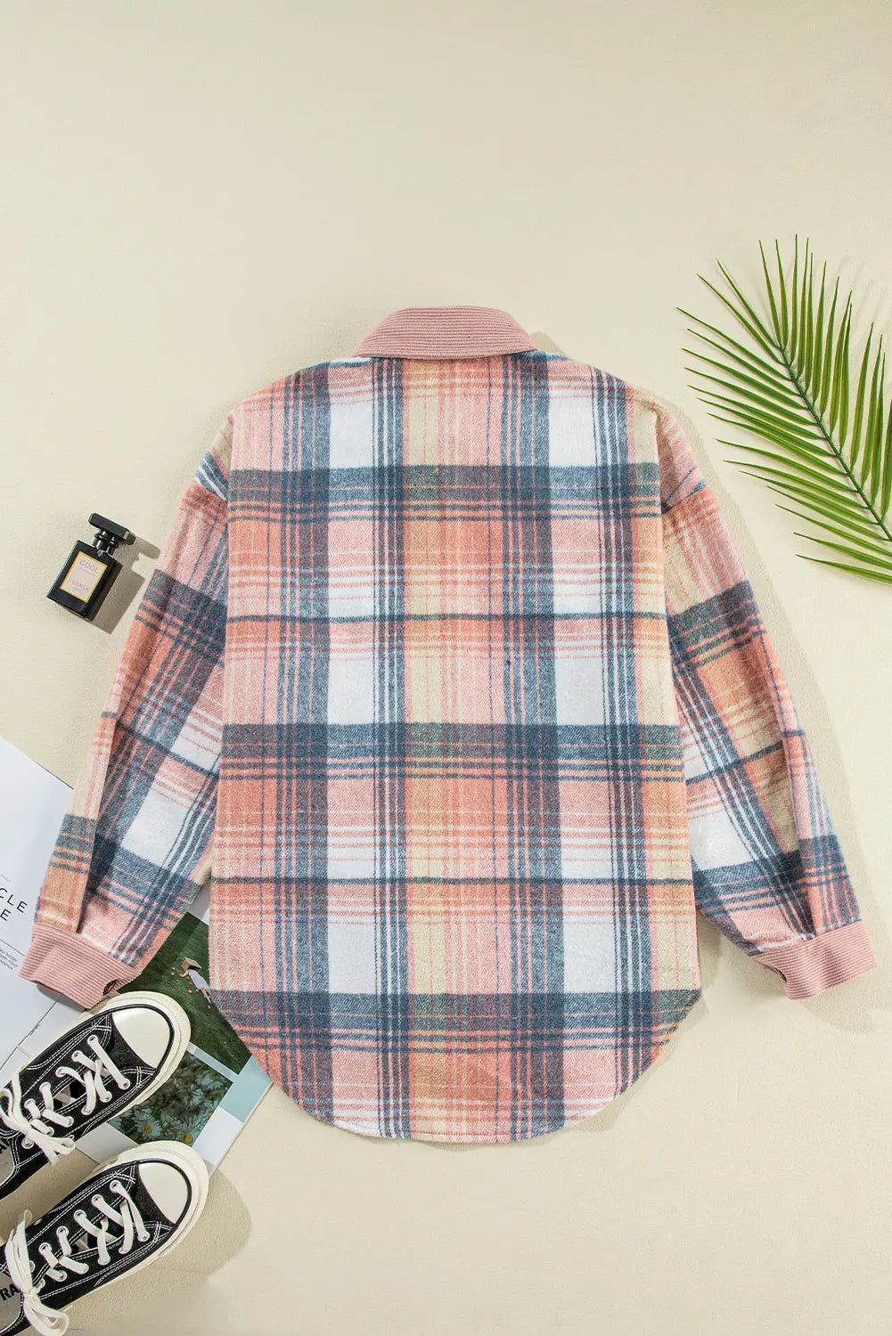 A long-sleeve plaid button-up shirt laid flat on a light beige background, styled with black sneakers, a perfume bottle, a green palm leaf, and a magazine. The shirt features a pink, blue, and white plaid pattern.