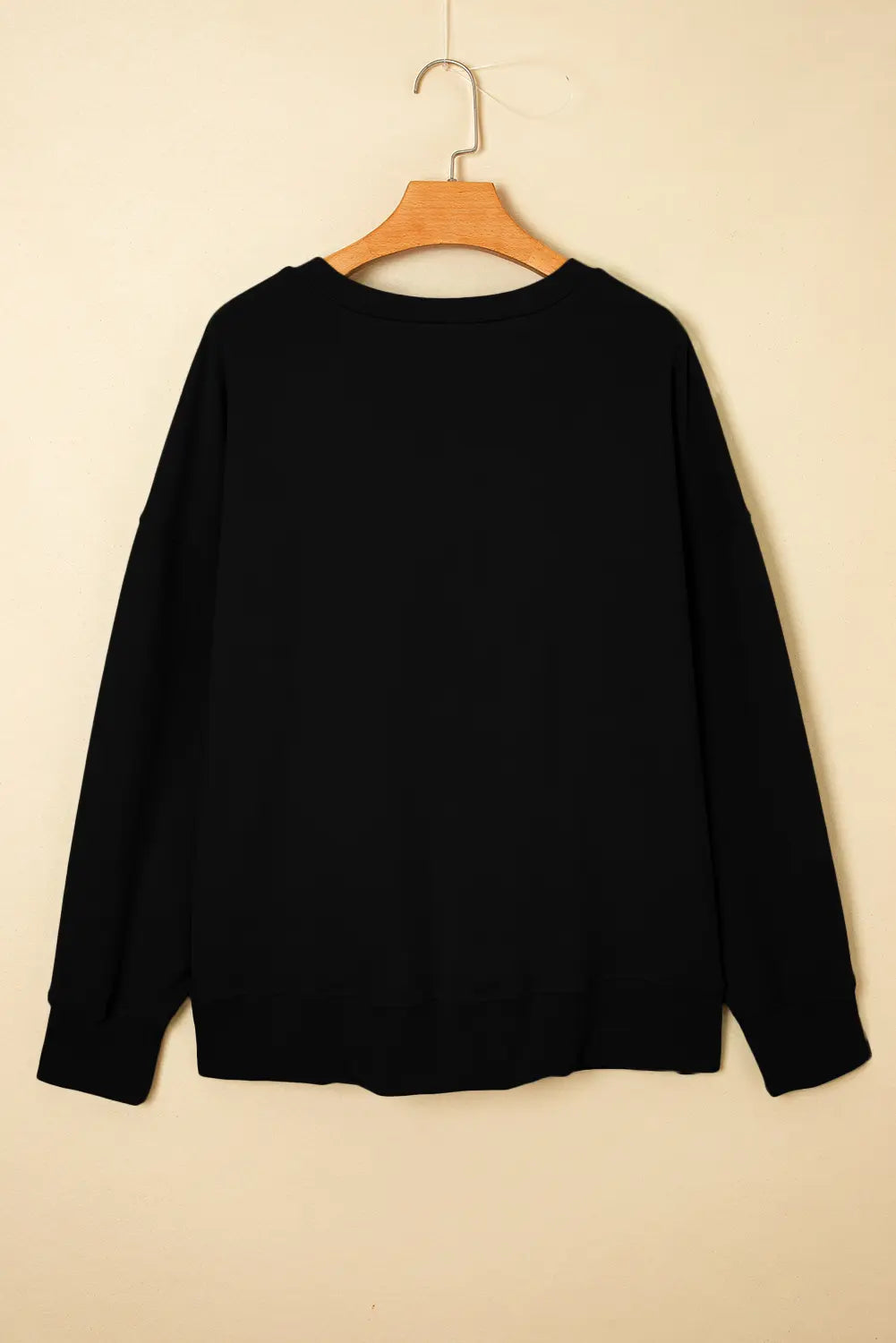 A black long-sleeve crewneck sweatshirt hanging on a wooden hanger against a plain beige wall.