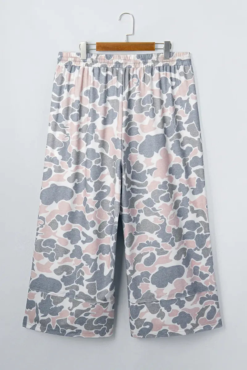 A pair of wide-leg cropped pants with an elastic waistband, featuring a pink, gray, and white camouflage pattern, hanging on a wooden hanger against a light gray wall.