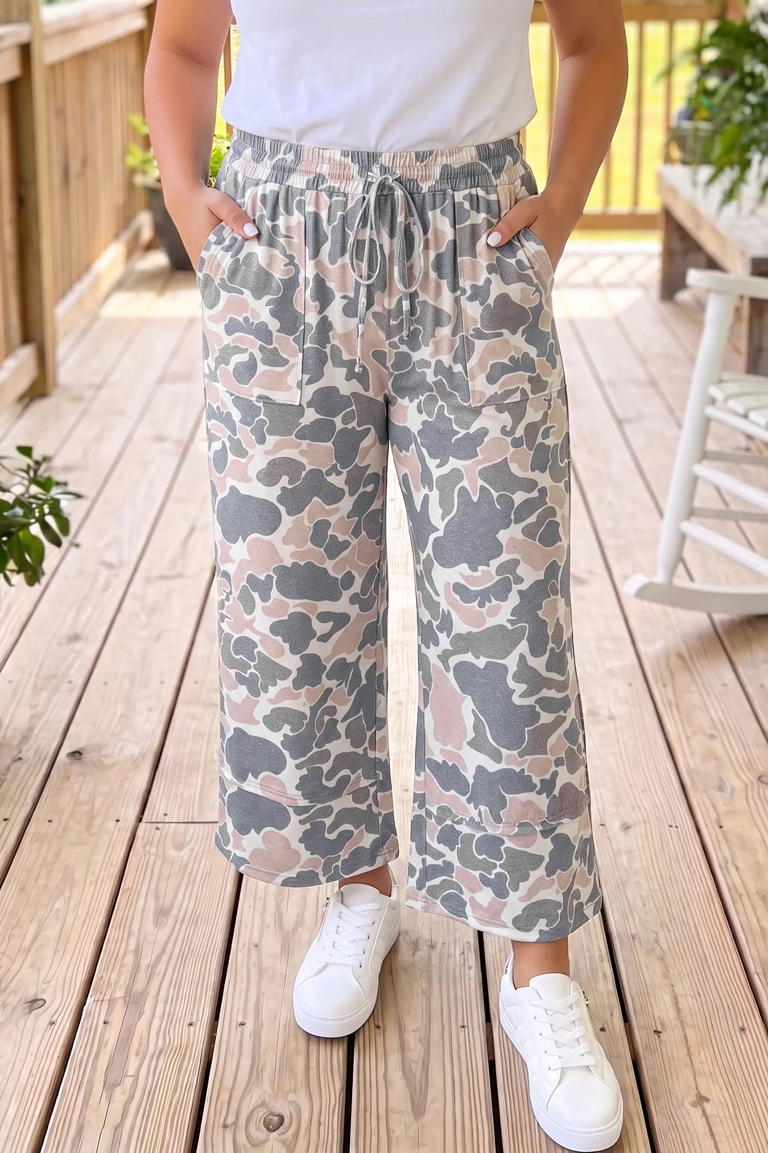 A person wearing loose, wide-leg drawstring pants with a gray, beige, and dusty pink camouflage pattern, paired with a white t-shirt and white sneakers, standing on a wooden deck.