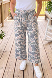 A person wearing loose, wide-leg drawstring pants with a gray, beige, and dusty pink camouflage pattern, paired with a white t-shirt and white sneakers, standing on a wooden deck.