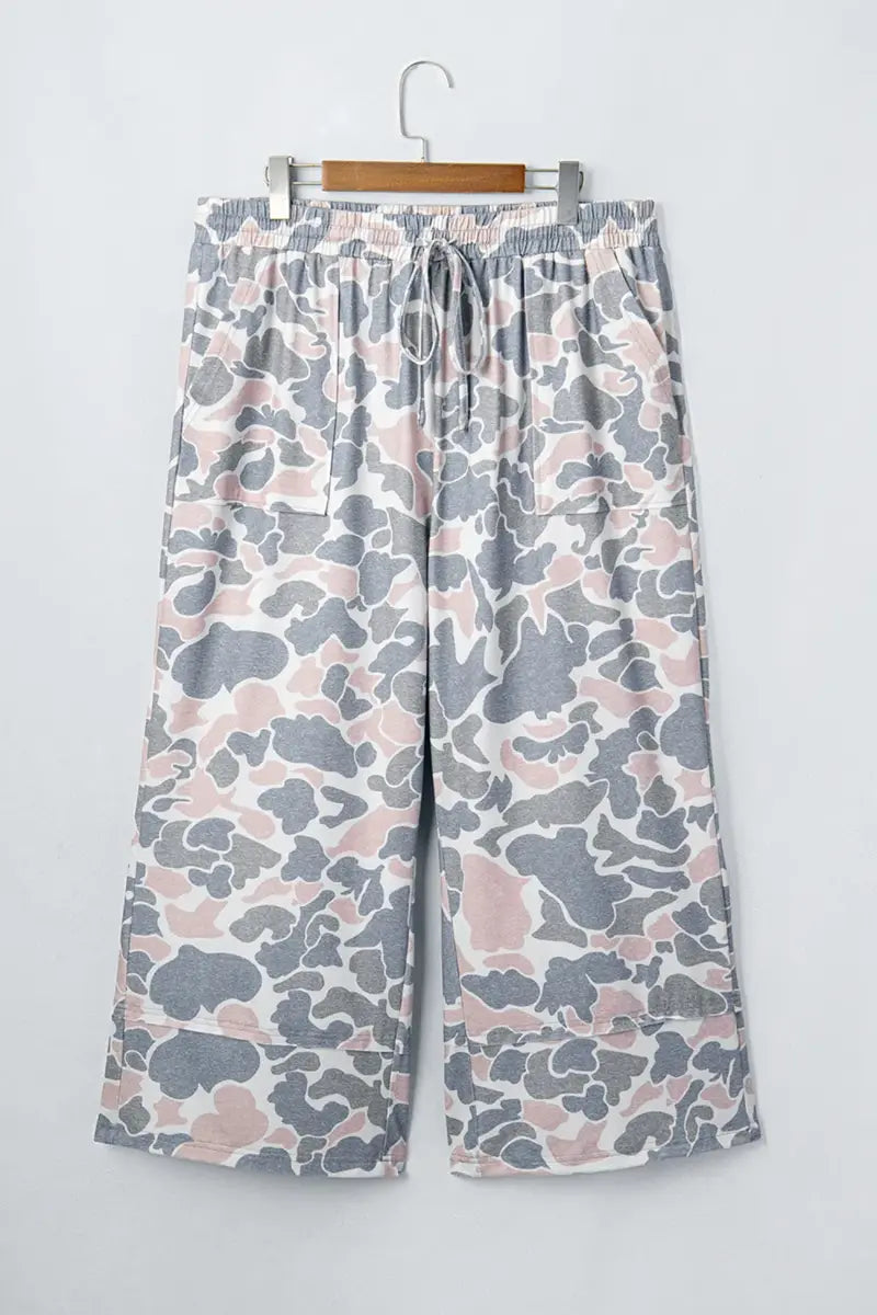 Wide-leg cropped pants with a gray, white, and dusty pink camouflage pattern, hanging on a wooden hanger against a light gray wall.