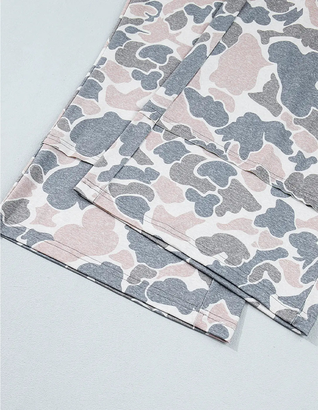 A close-up of folded fabric with an abstract organic camouflage-like pattern in soft pink, gray, and off-white on a light gray background.
