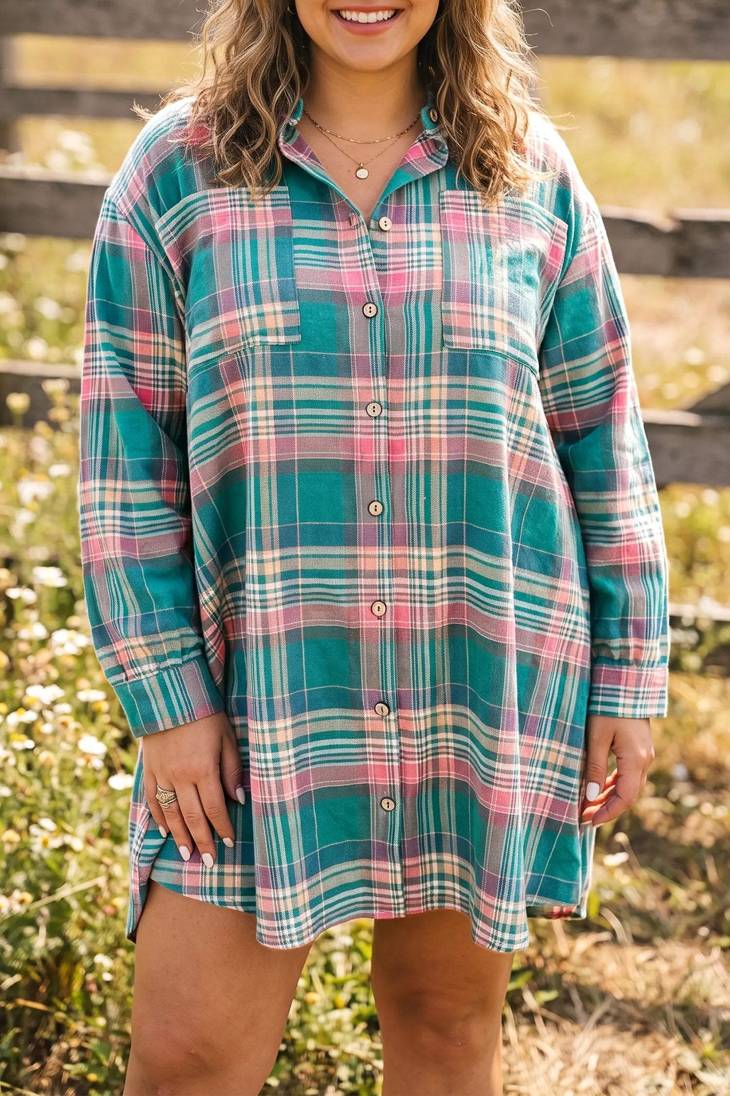 A woman wearing a long-sleeve plaid button-up shirt dress in teal green, pink, and white, standing outdoors in a grassy field.