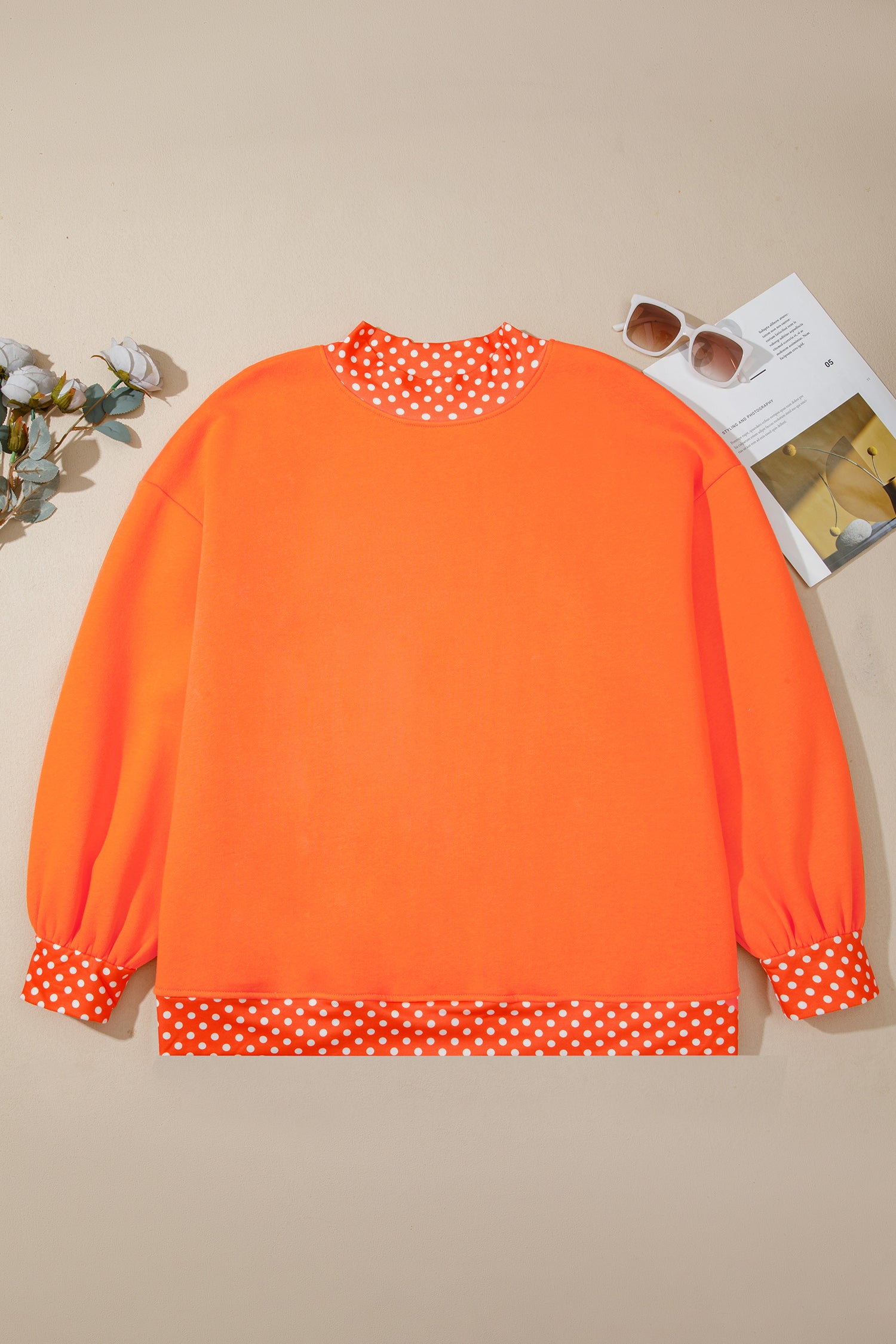Bright orange long-sleeve sweatshirt with white polka dot trim at the crew neck, cuffs, and hem, styled flat on a beige background with sunglasses and flowers.