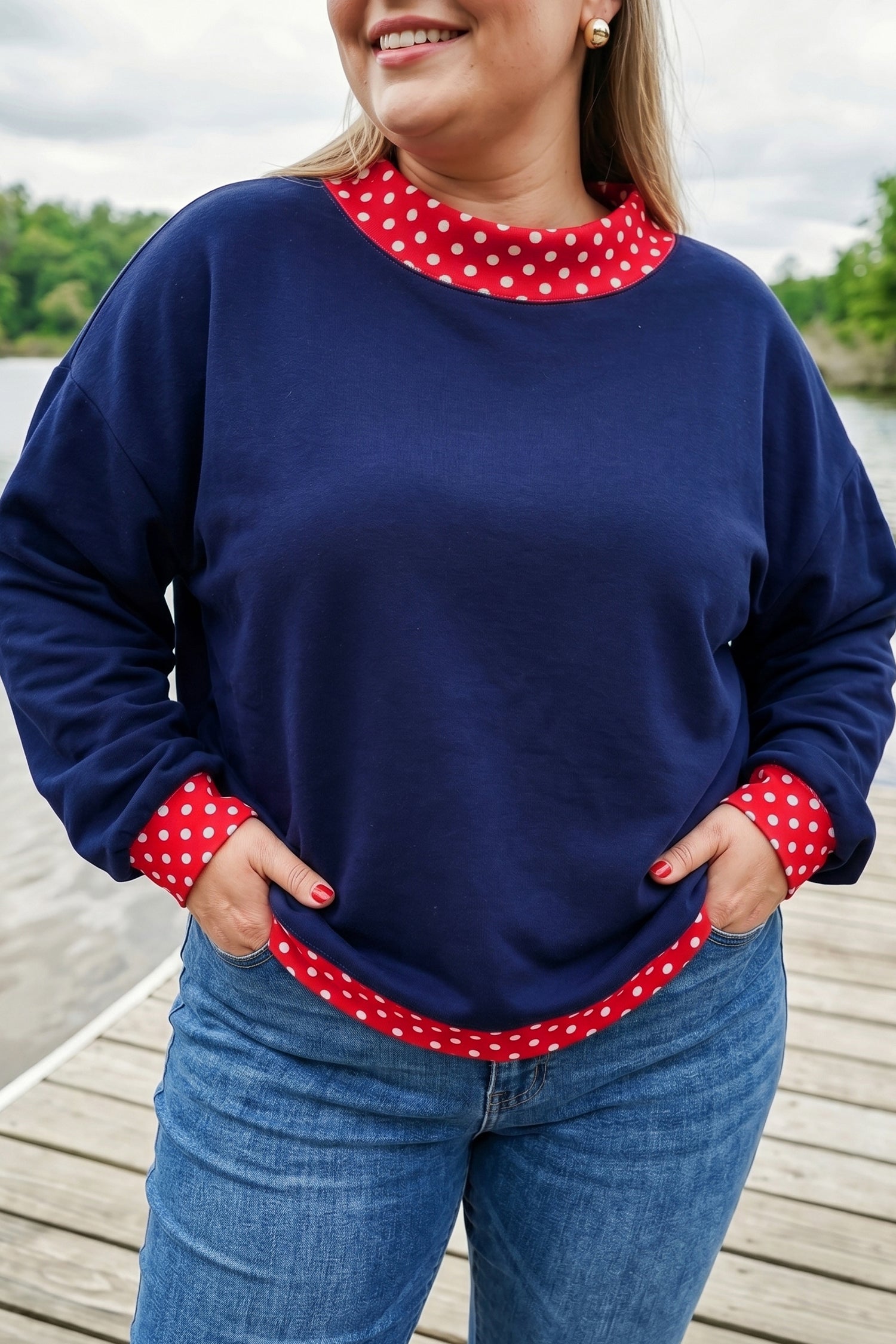 A woman wearing a navy blue long-sleeve sweatshirt with red and white polka dot trim at the crew neck, cuffs, and hem, paired with blue jeans, standing on a wooden dock.