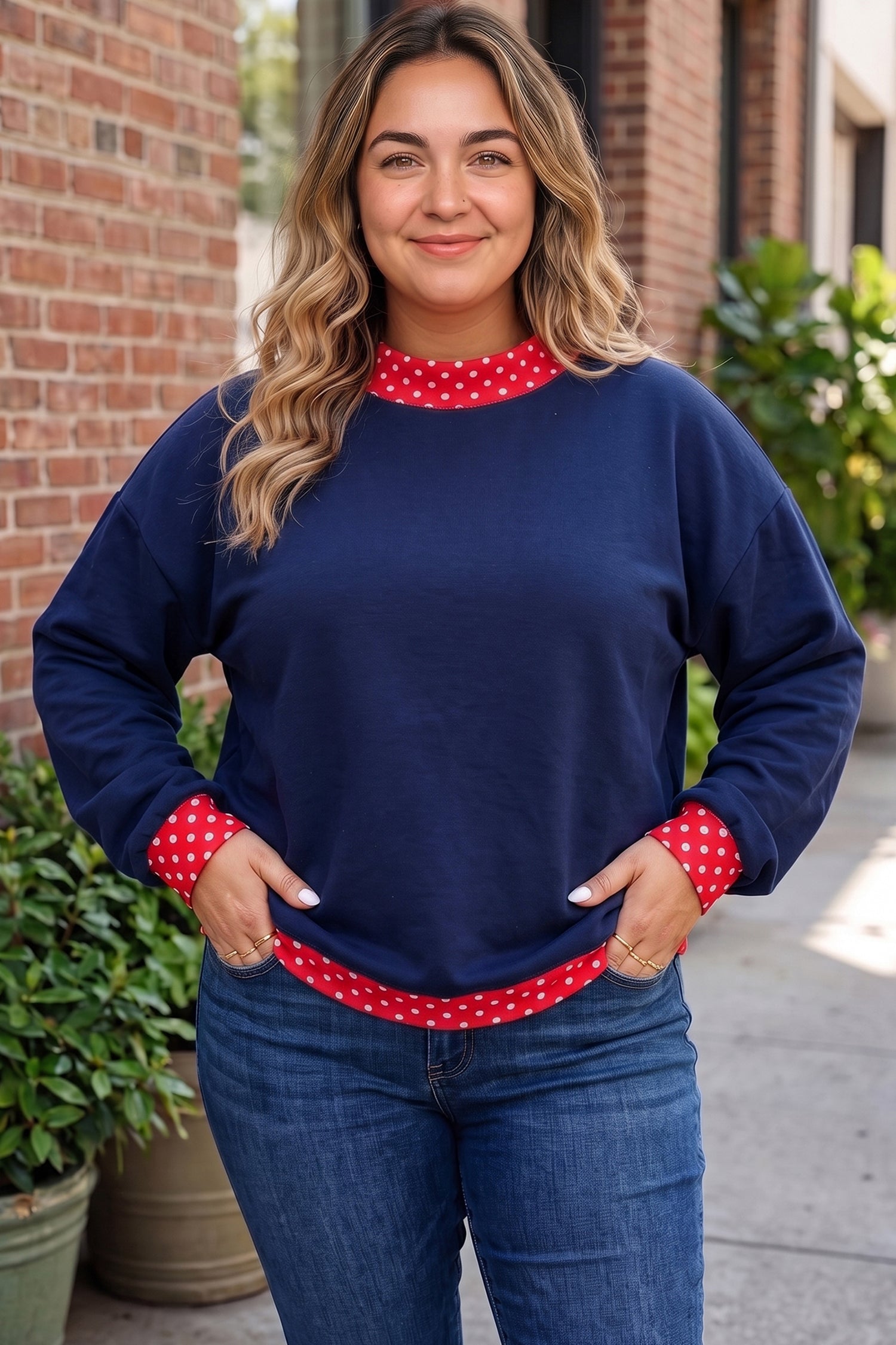 A woman wearing a navy blue crewneck sweatshirt with red and white polka dot trim at the collar, cuffs, and hem, paired with blue jeans, standing outdoors in front of a brick building.