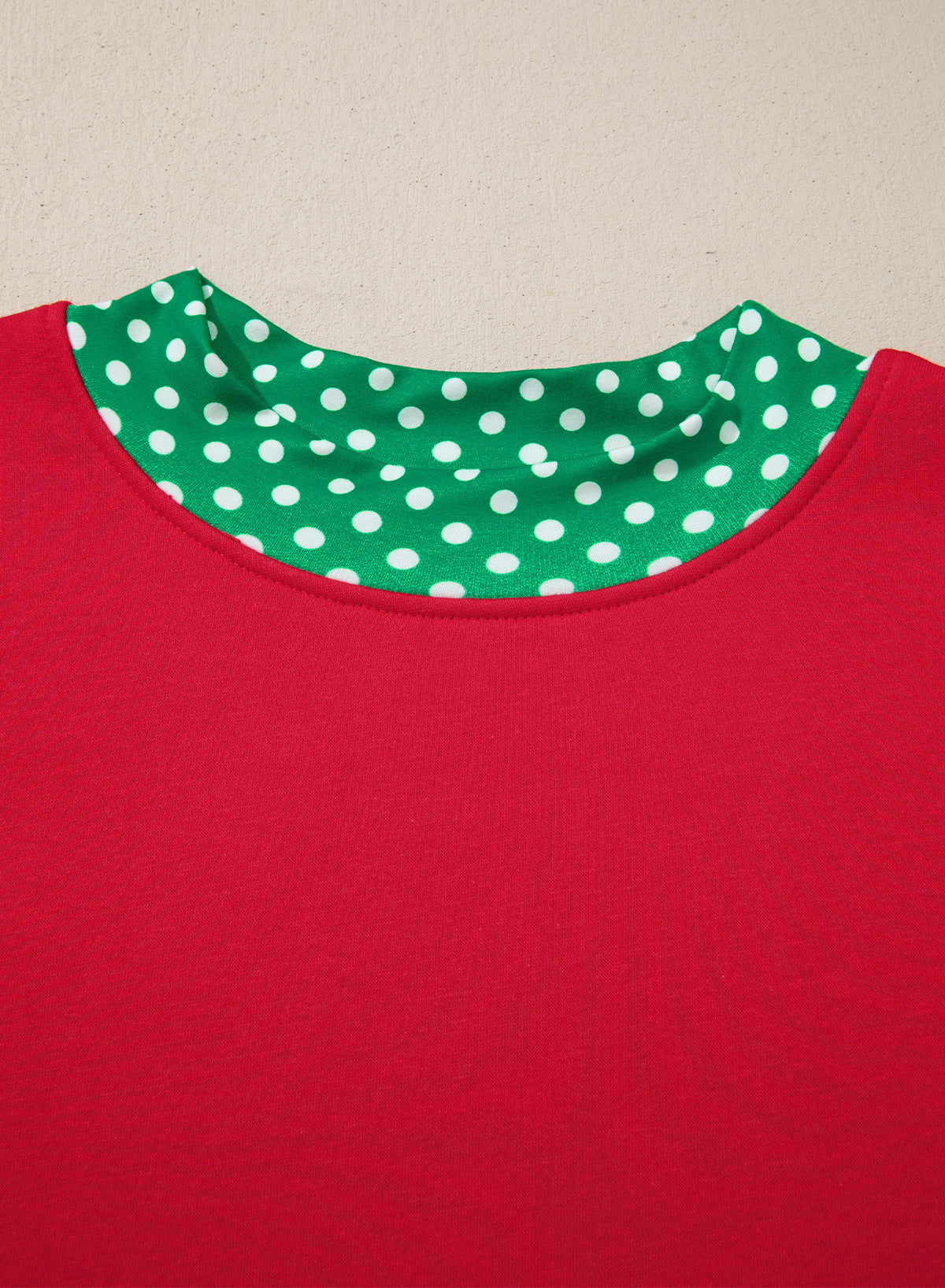 Close-up of a red garment with a contrasting green and white polka dot inner collar, shown against a plain beige background.
