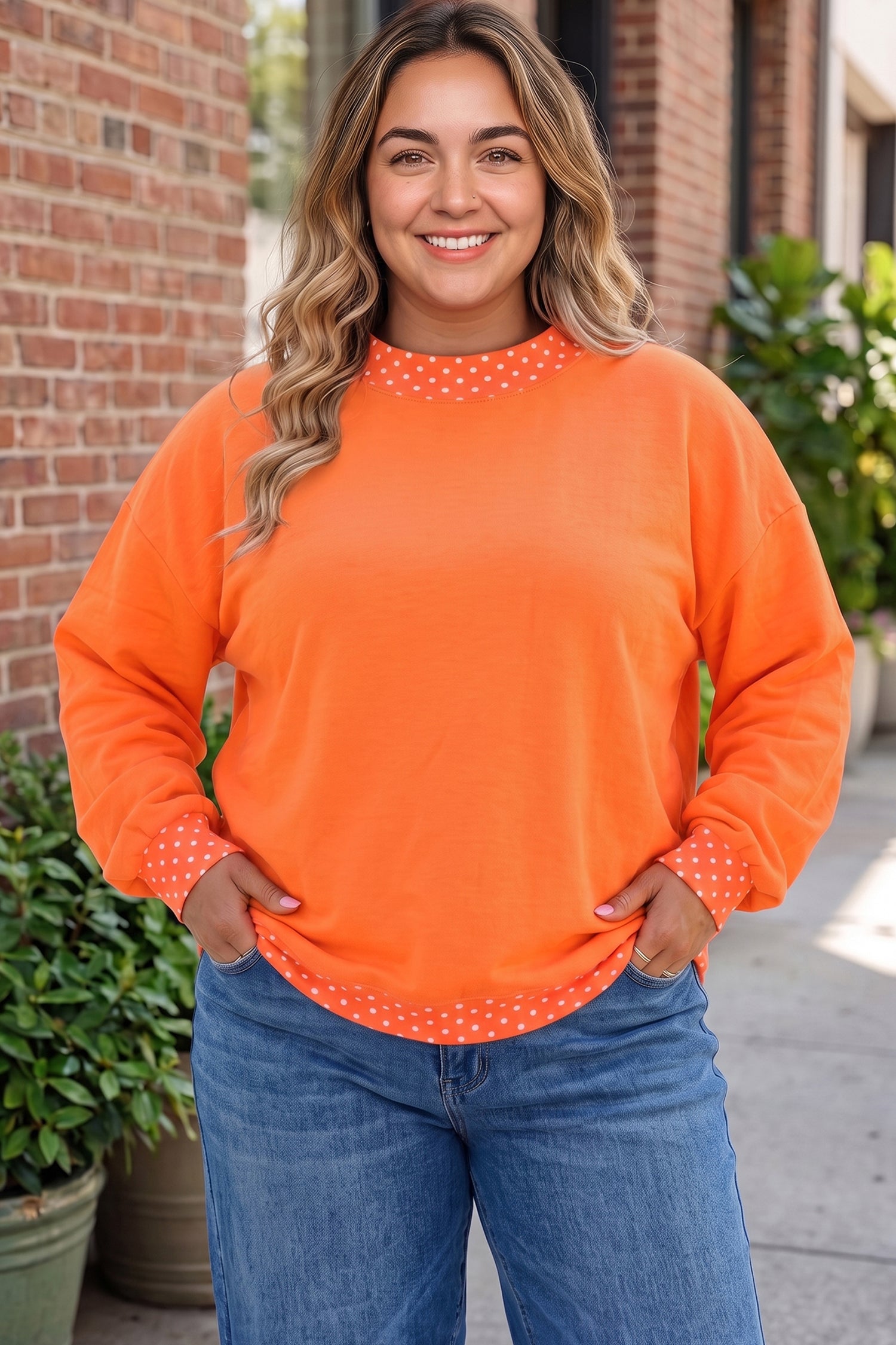 A woman wearing a bright orange long-sleeve crewneck sweatshirt with white polka dot trim around the collar and cuffs, paired with blue jeans, standing outdoors in front of a brick building.