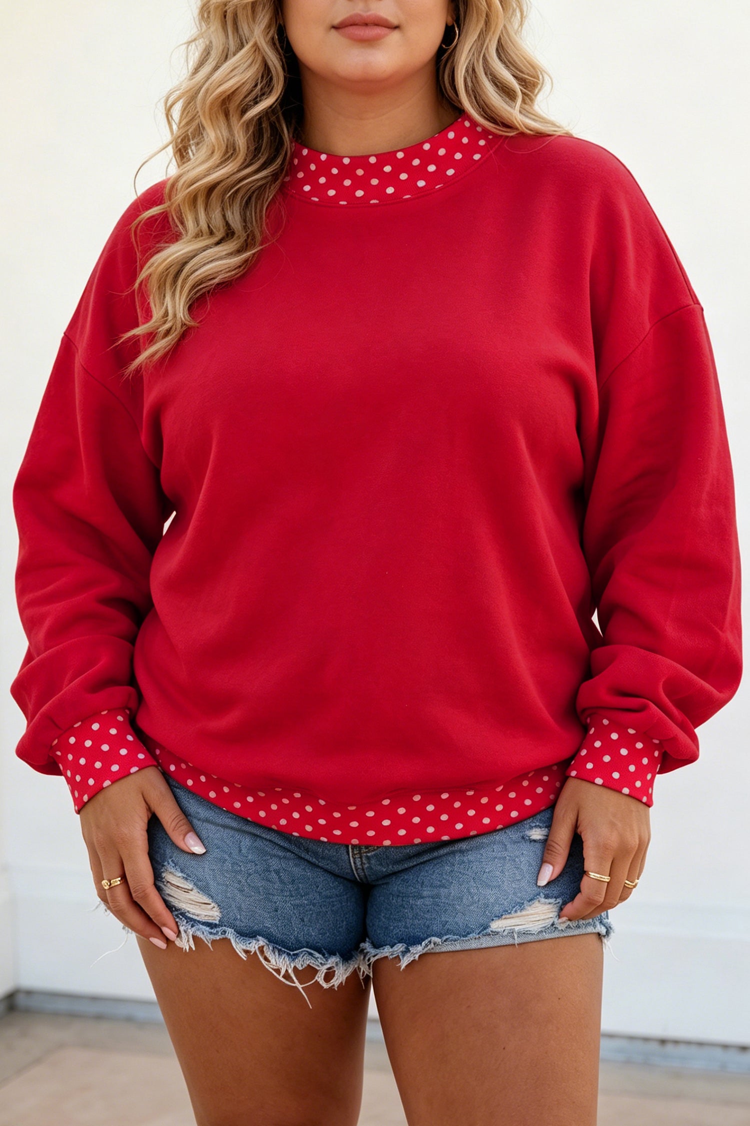 A woman wearing a red crewneck sweatshirt with white polka dot trim at the collar, cuffs, and hem, paired with distressed denim shorts.