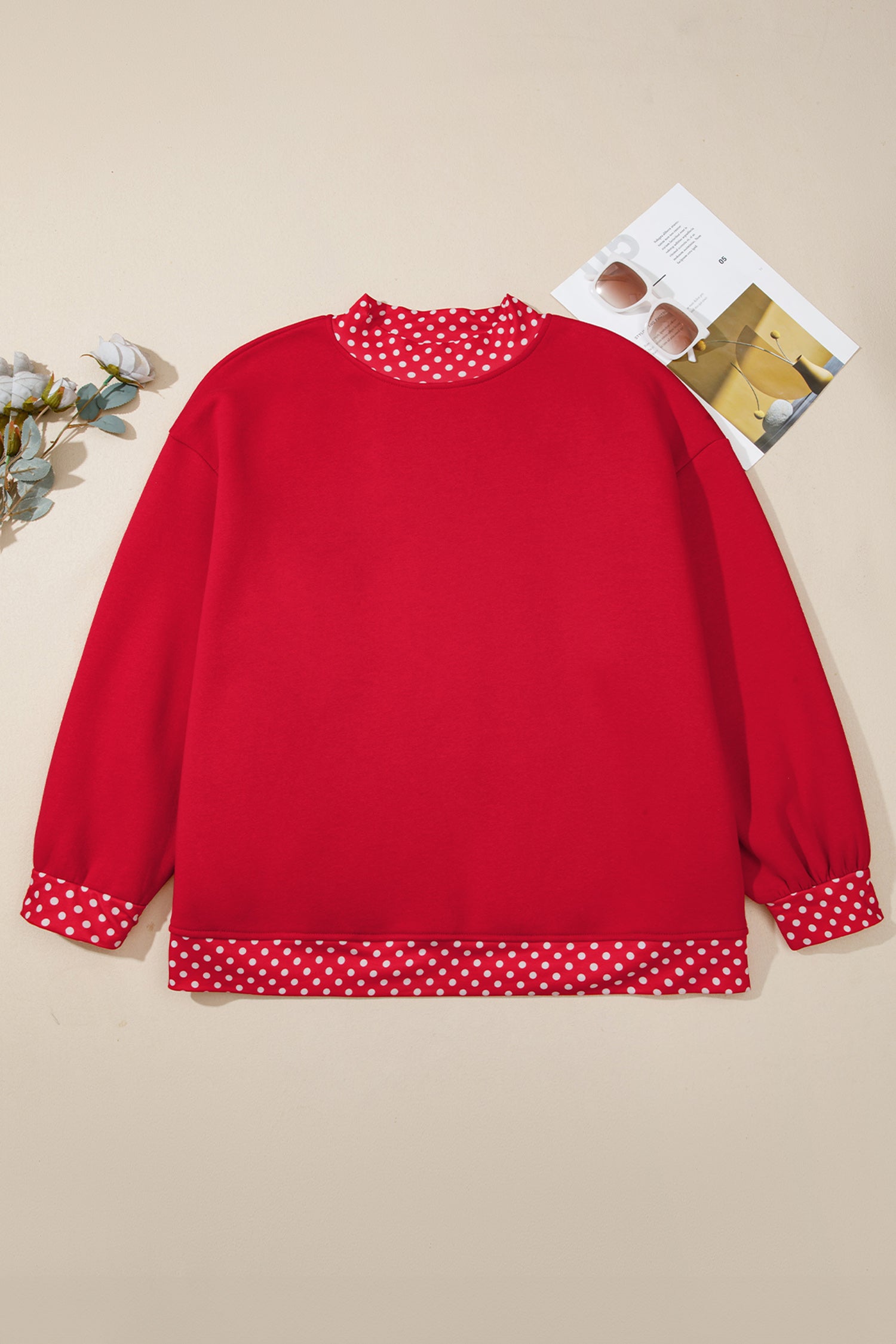 A red long-sleeve sweatshirt with red and white polka dot trim on the crew neck, cuffs, and hem, displayed flat on a beige background with a small decorative card and flowers.