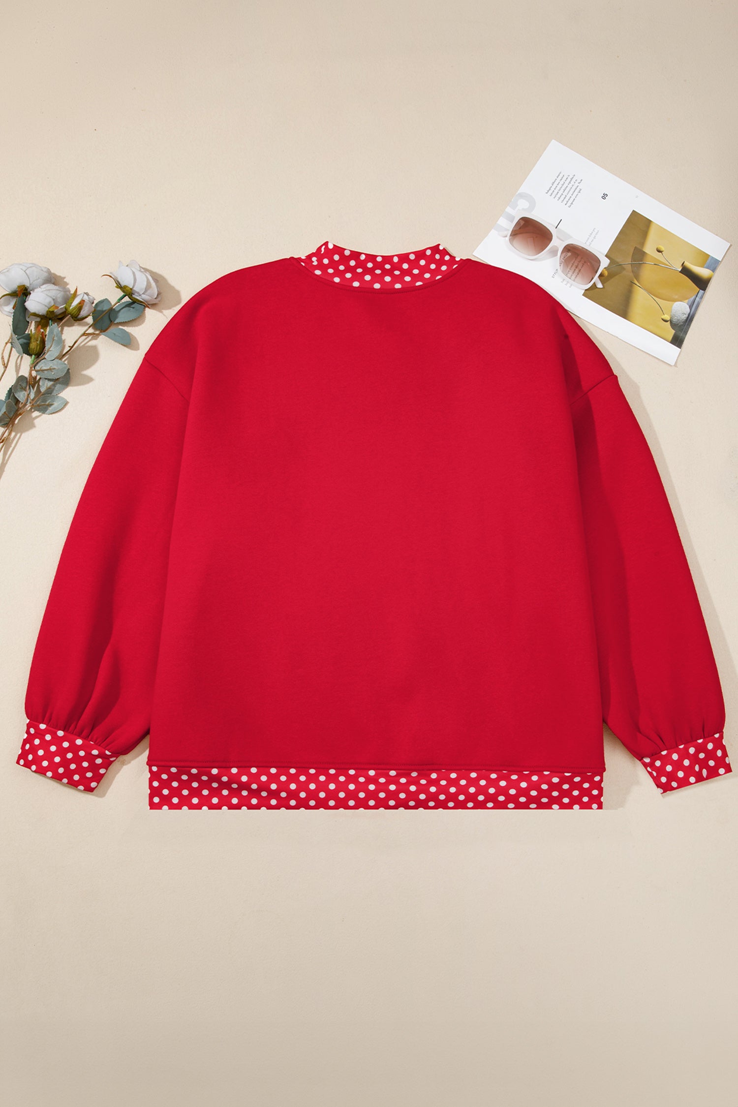A red long-sleeve sweatshirt with red and white polka dot trim on the collar, cuffs, and hem, laid flat on a beige background with decorative flowers and a magazine.