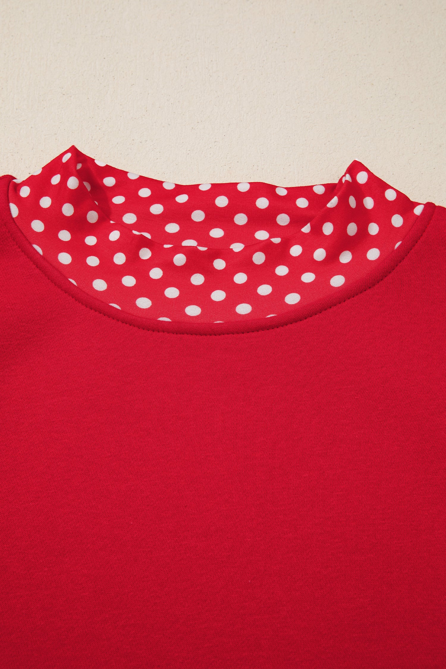 Close-up of a red crewneck top with a white polka-dot patterned inner collar lining, set against a plain light beige background.