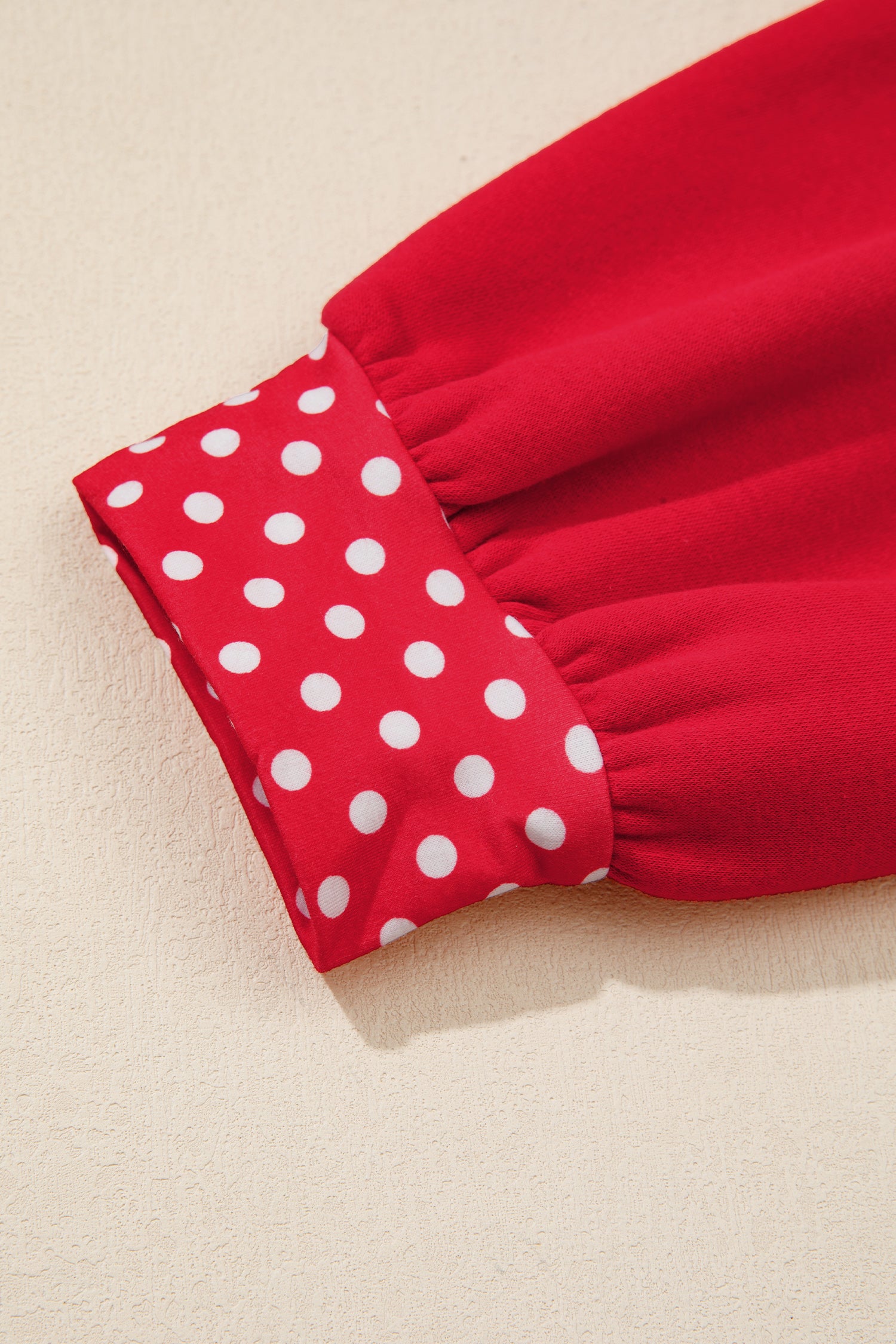 Close-up of a red garment sleeve with a gathered cuff featuring a white polka dot pattern, set against a light beige textured background.