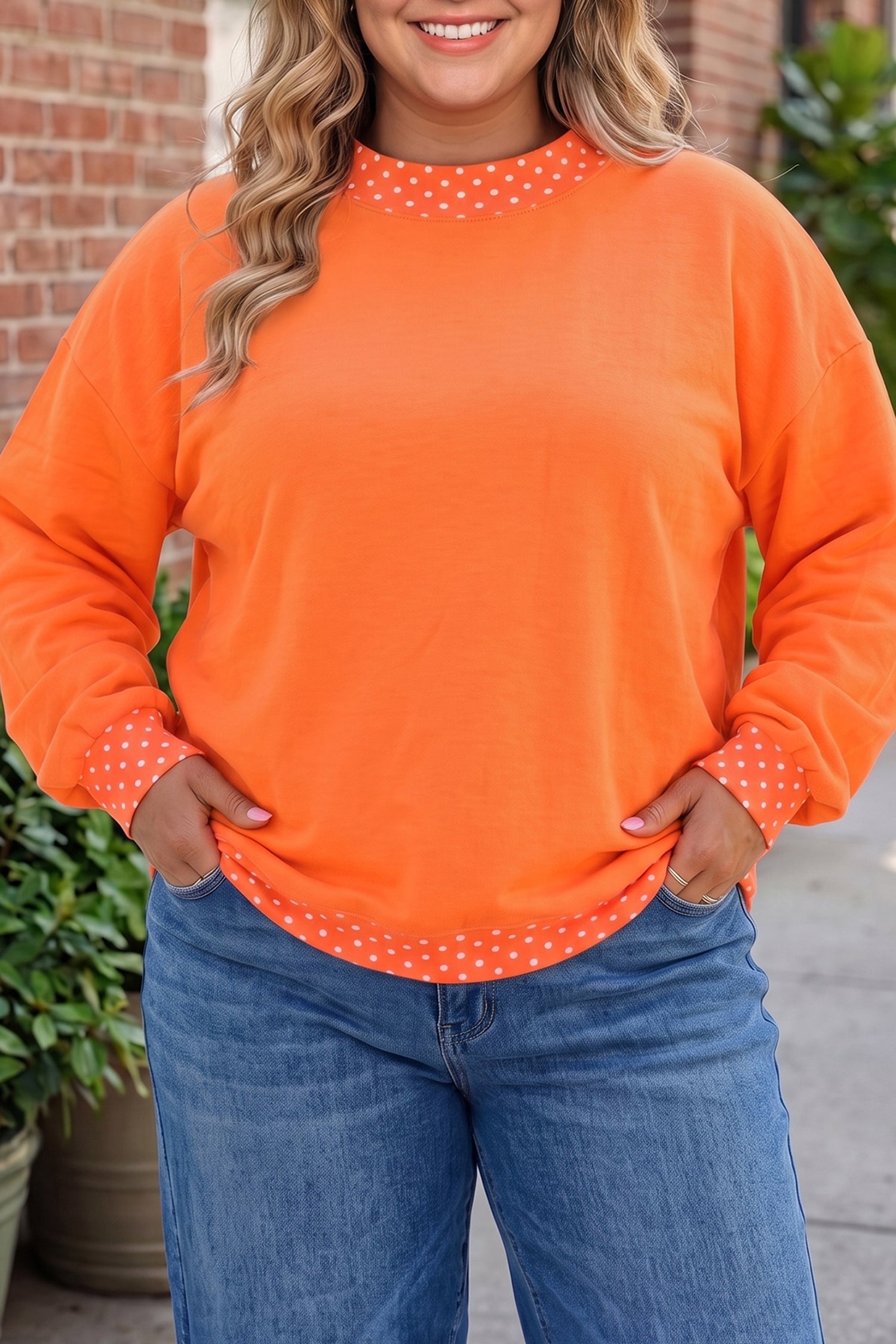 A woman wearing a bright orange long-sleeve crewneck sweatshirt with white polka dot trim around the collar, cuffs, and hem, paired with blue jeans.