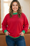 A woman wearing a festive red crewneck sweater with green and white polka dot accents on the collar and cuffs, paired with blue jeans.