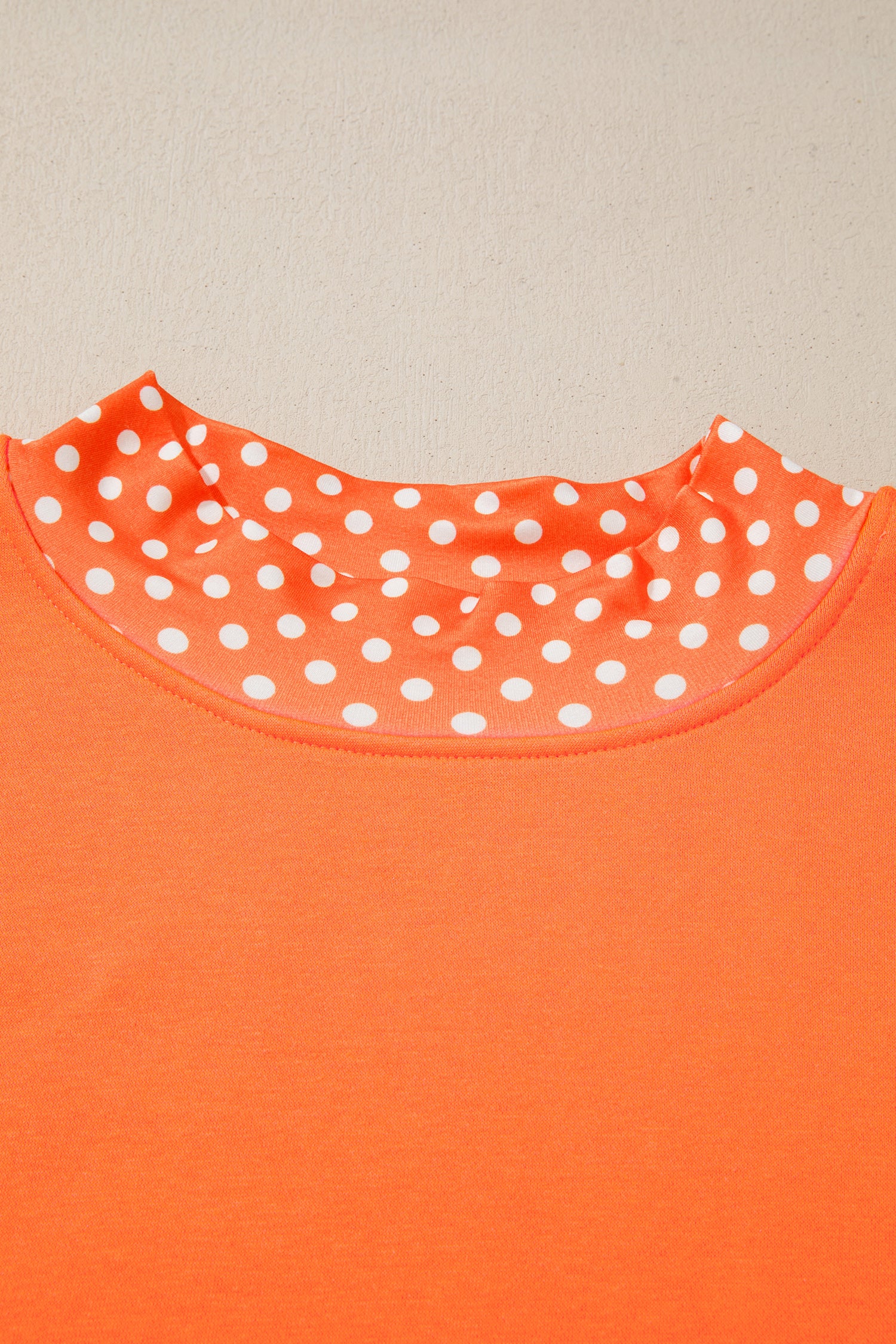 Close-up of a bright orange top with a white polka dot pattern lining the inner neckline, set against a plain light beige background.