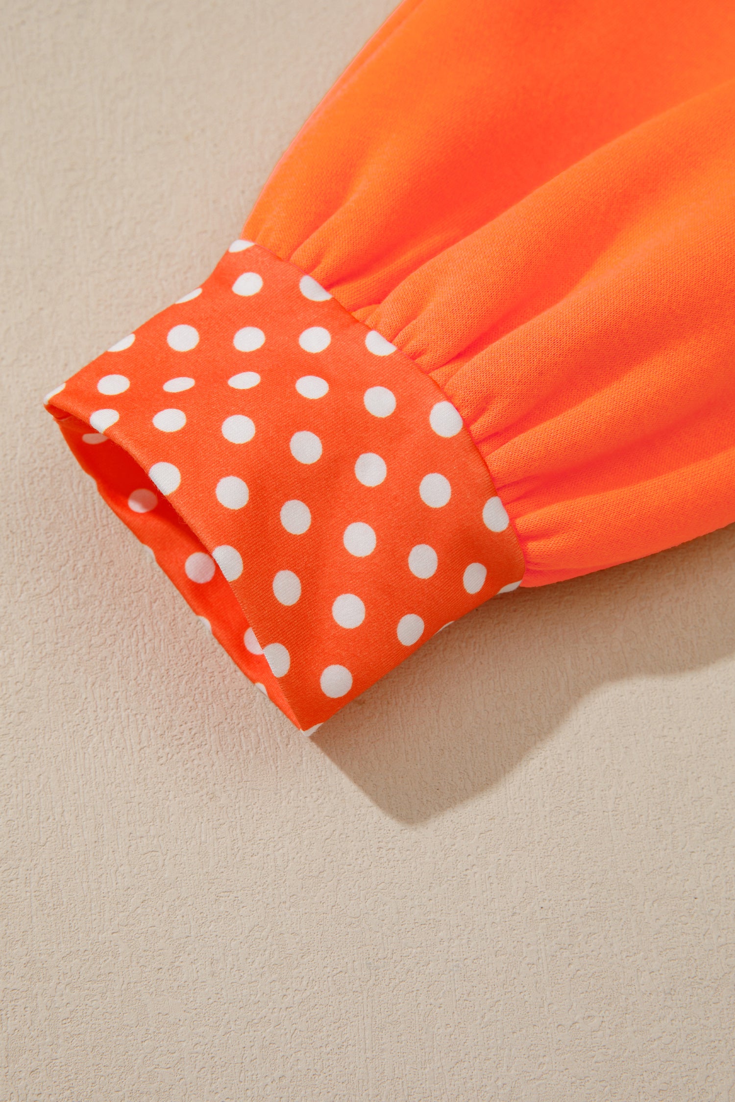 Close-up of an orange garment sleeve with a gathered cuff featuring a white polka dot pattern, set against a neutral beige background.