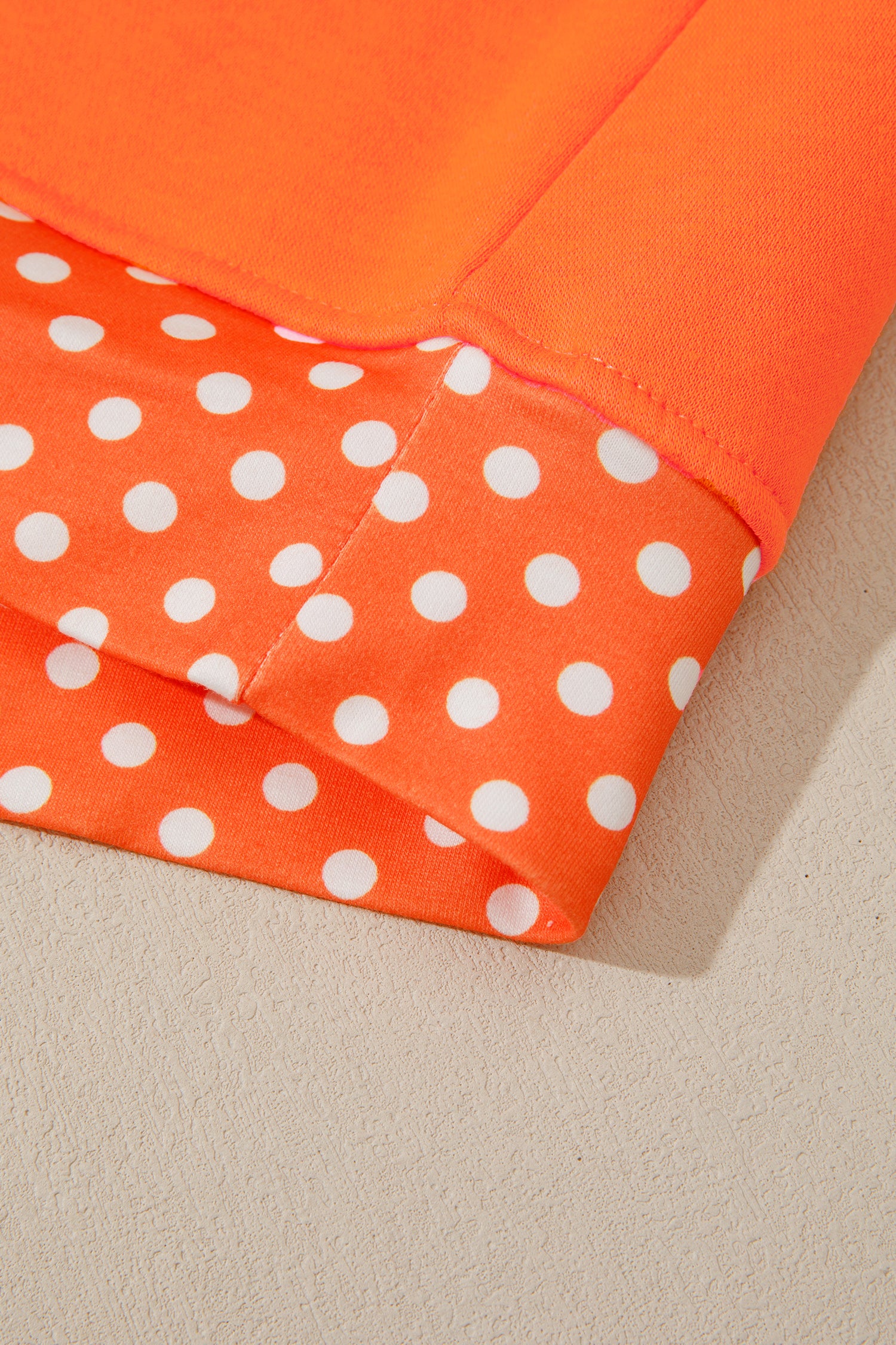 Close-up of folded fabric in bright orange, including one section with white polka dots and a solid orange reverse side, laid on a light beige textured surface.