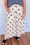 A person wearing cream wide-leg pants with large brown polka dots, side pockets, and an elastic waistband, paired with a slate blue sleeveless top and black slide sandals, standing against a white paneled wall and wood floor.