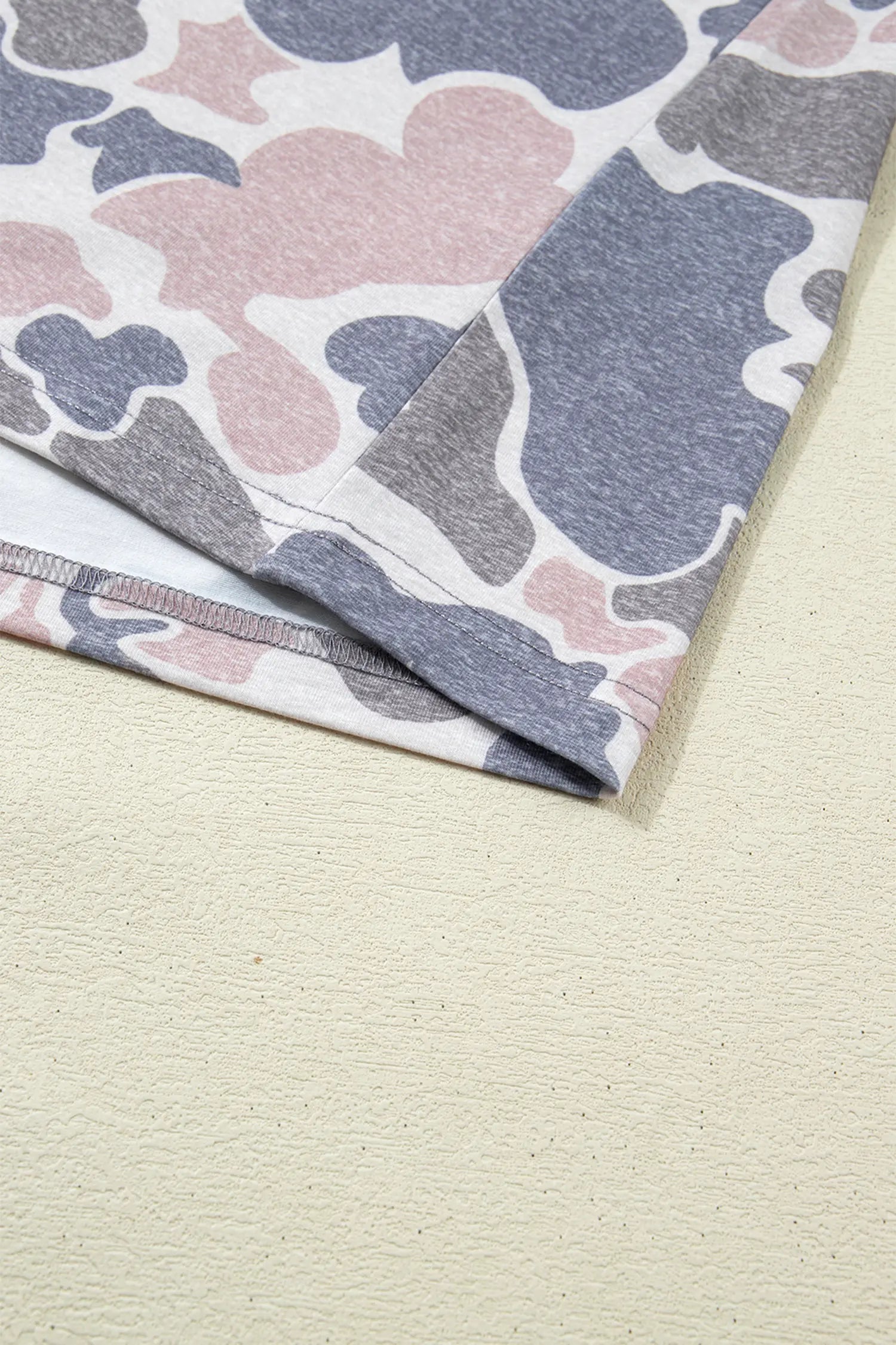A close-up of a folded fabric item, likely a sweatshirt or blanket, featuring a pastel camouflage pattern in pink, slate blue, and gray on a white base, set against a textured beige background.