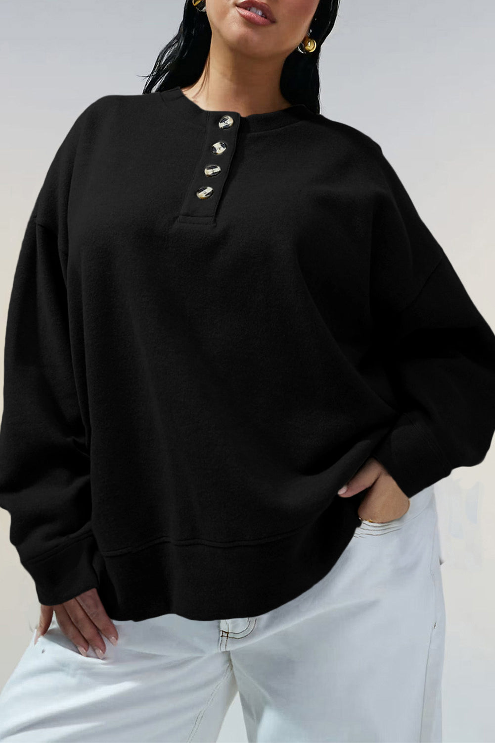 Woman wearing an oversized black henley sweatshirt with gold-tone buttons and white pants.