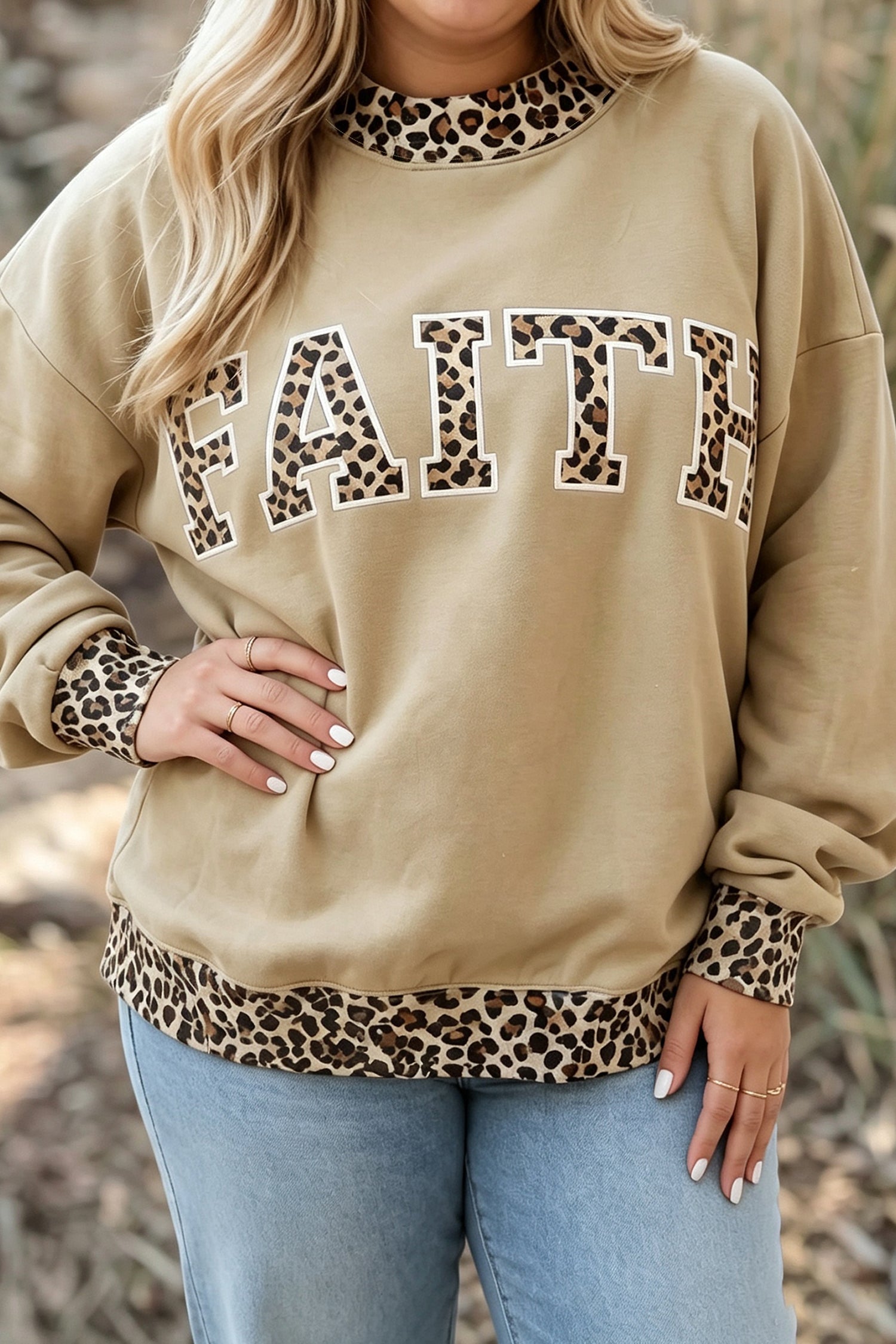 Woman wearing a beige sweatshirt with leopard trim and “FAITH” lettering, paired with light blue jeans.