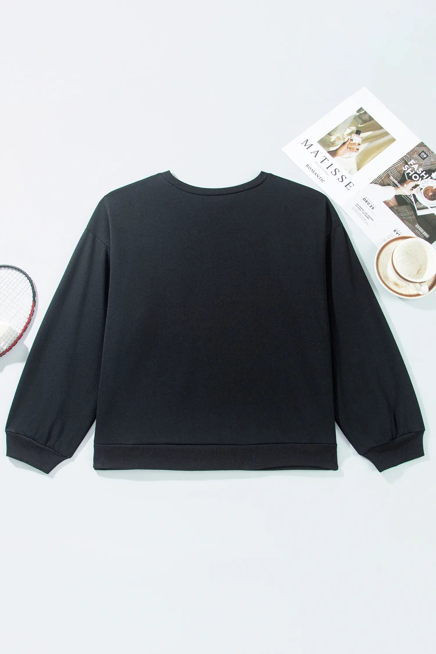 Black long-sleeve crewneck sweatshirt laid flat with a magazine, cup, and tennis racket.