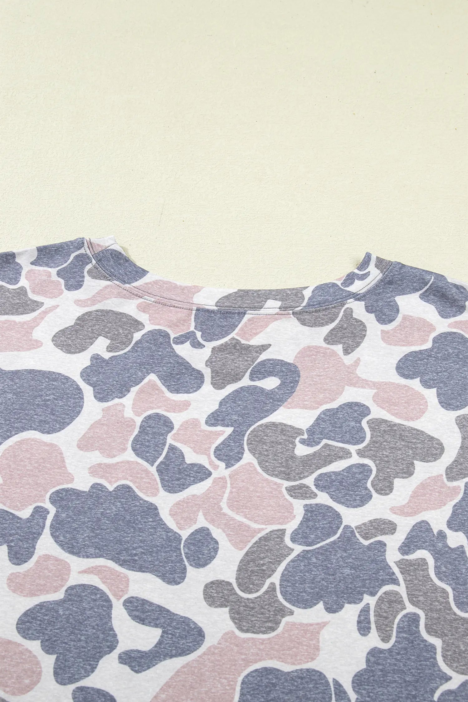 A close-up of a crewneck t-shirt featuring a pink, gray, and blue-gray abstract camouflage pattern on a white base, laid flat against a light beige background.