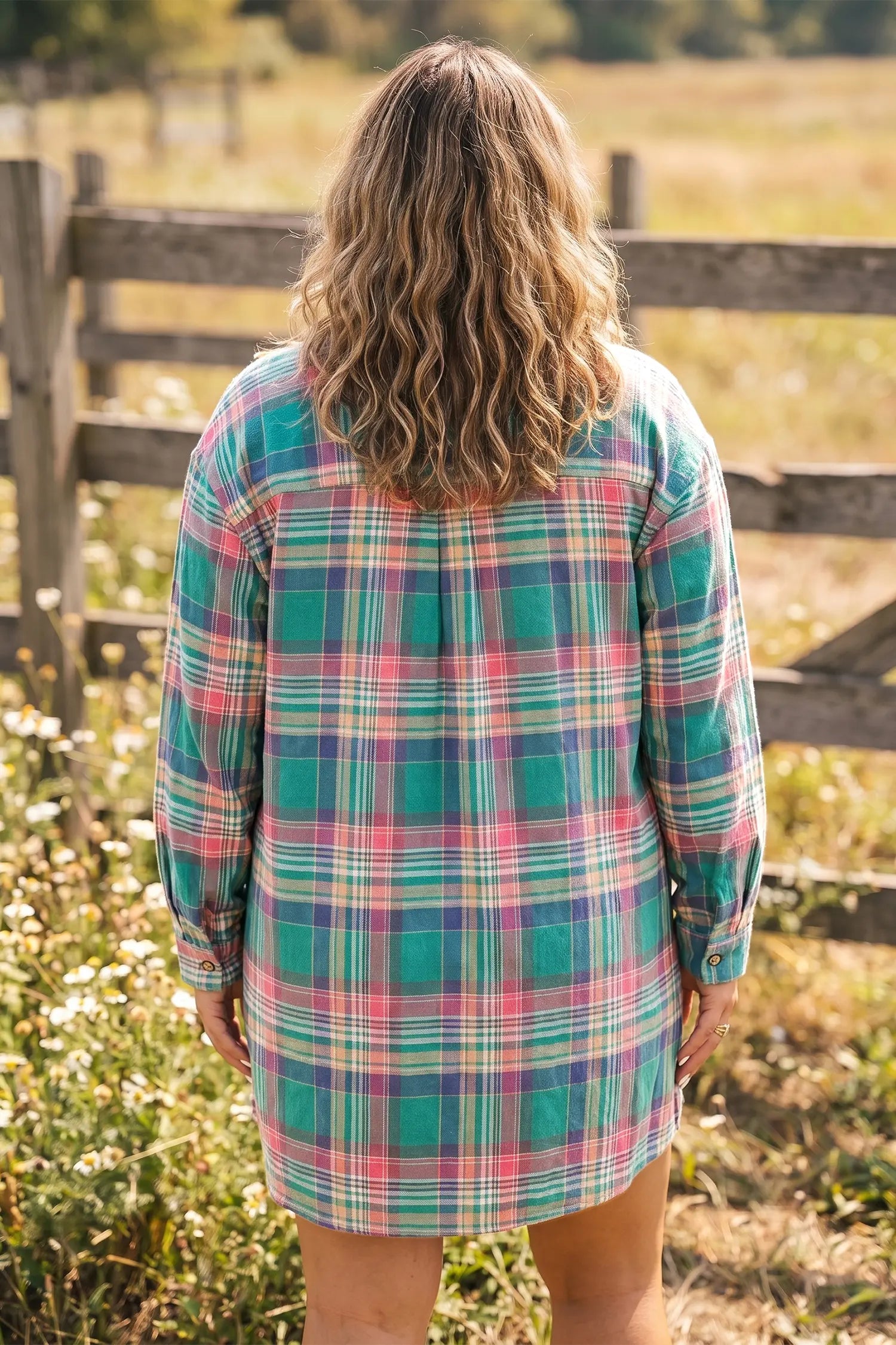 A woman viewed from behind wearing a long-sleeve plaid shirt dress in green, pink, and blue, standing outdoors in front of a wooden fence and grassy field.