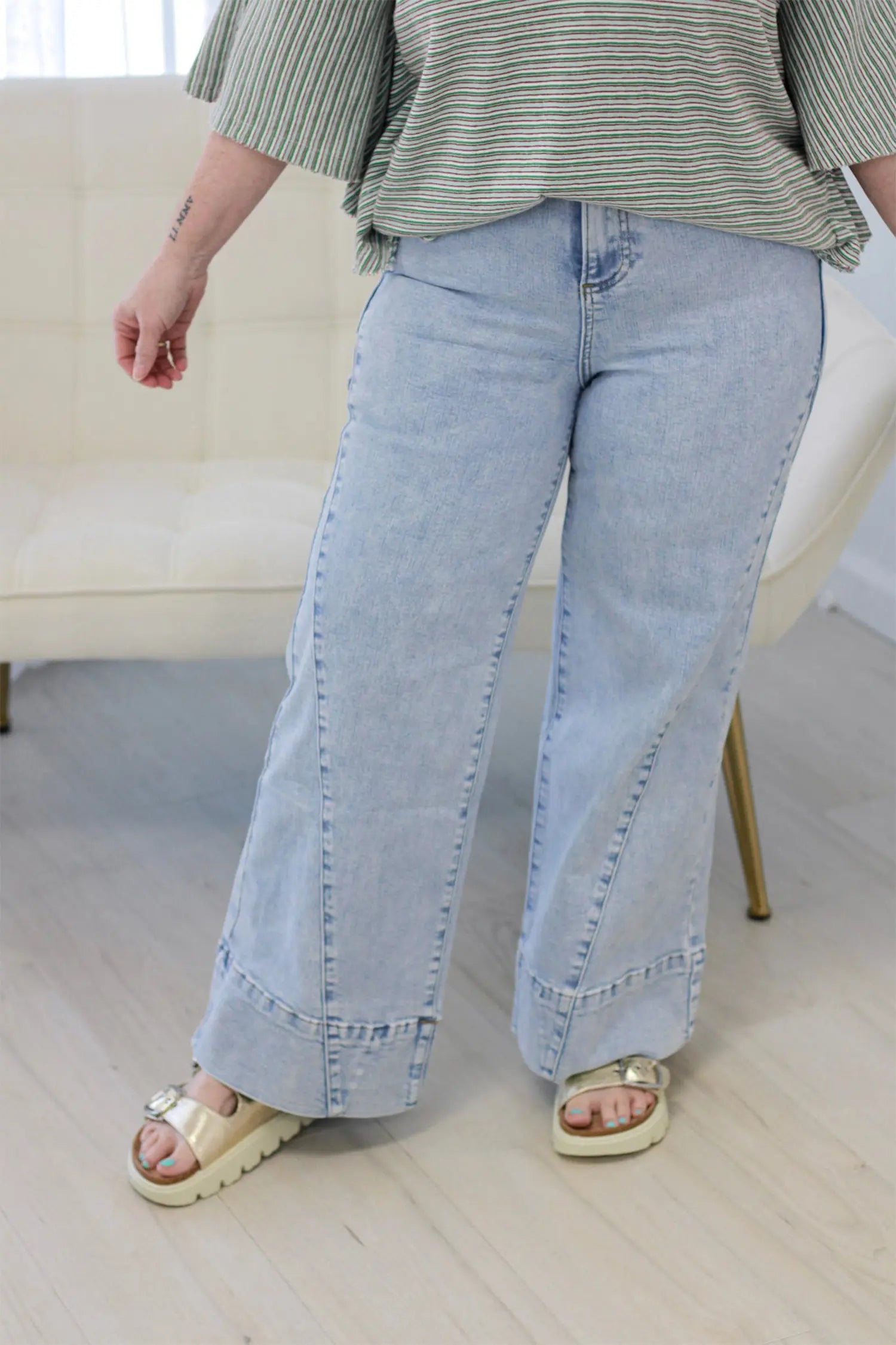 A person wearing light-wash wide-leg denim jeans with rolled cuffs, paired with a green striped top and beige platform sandals, standing in a bright indoor room.